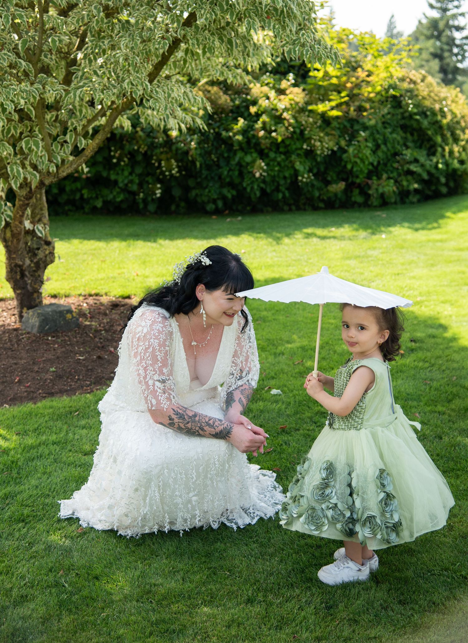 Bride in lace gown crouches to talk with young flower girl in green dress holding white parasol.