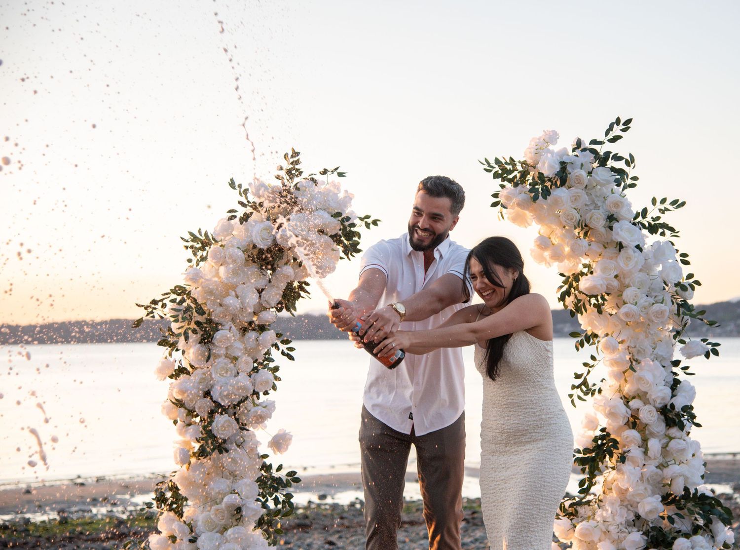 Couple celebrating engagement with champagne by floral arch on beach at sunset.
