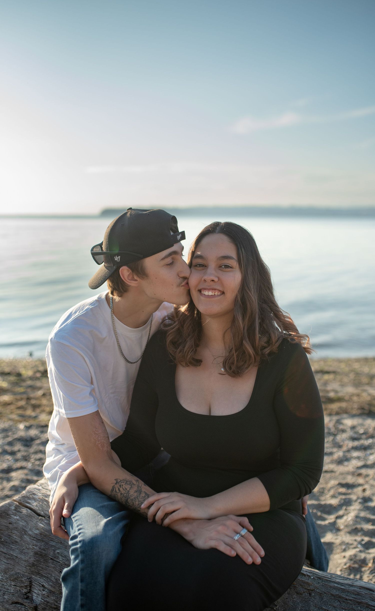 Couple sitting on driftwood at beach, man kissing woman's cheek as she smiles, ocean backdrop.