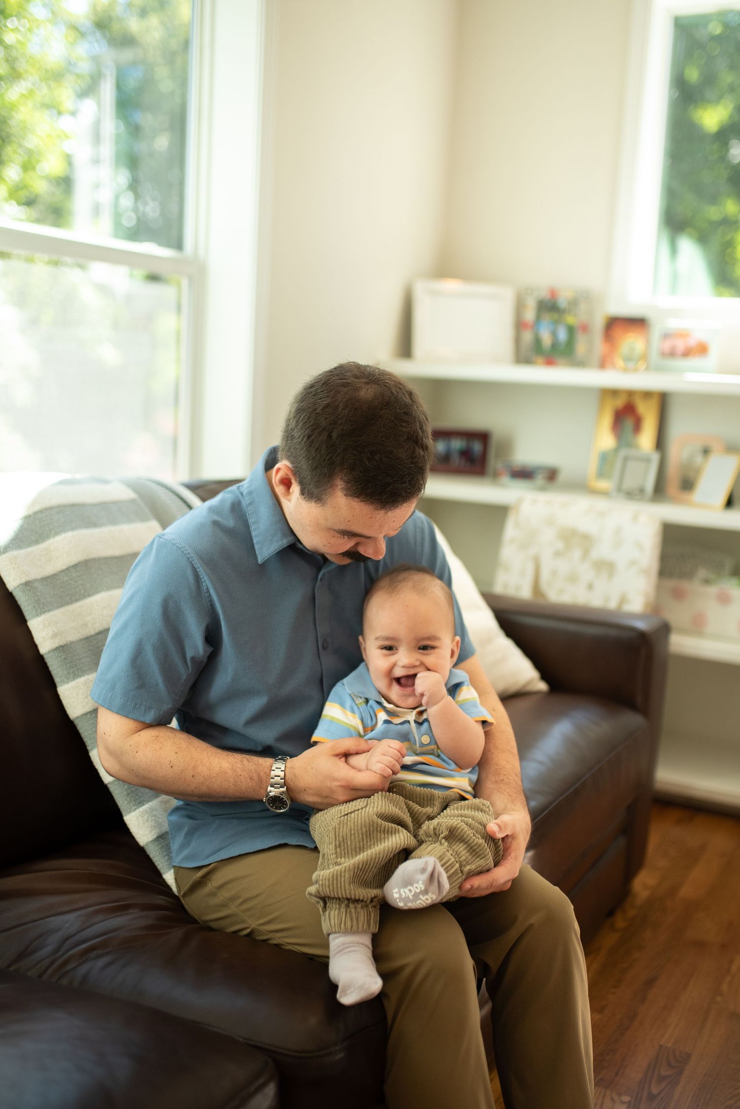 Father sitting on a leather sofa, laughing and bonding with his happy smiling baby boy in a bright living room.