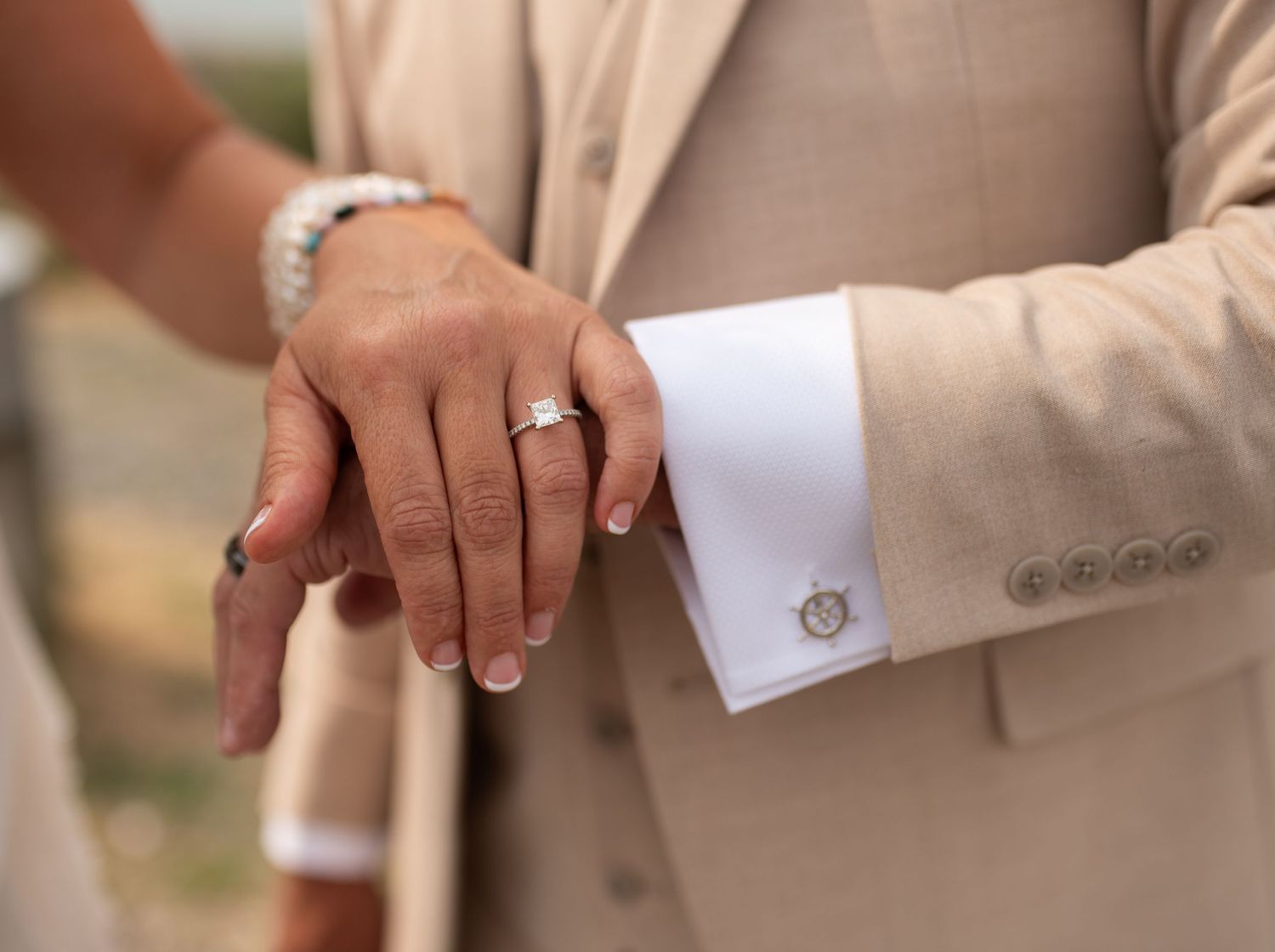 Close-up of a couple's hands touching, woman wearing a diamond engagement ring, man in a beige suit with silver cufflinks.