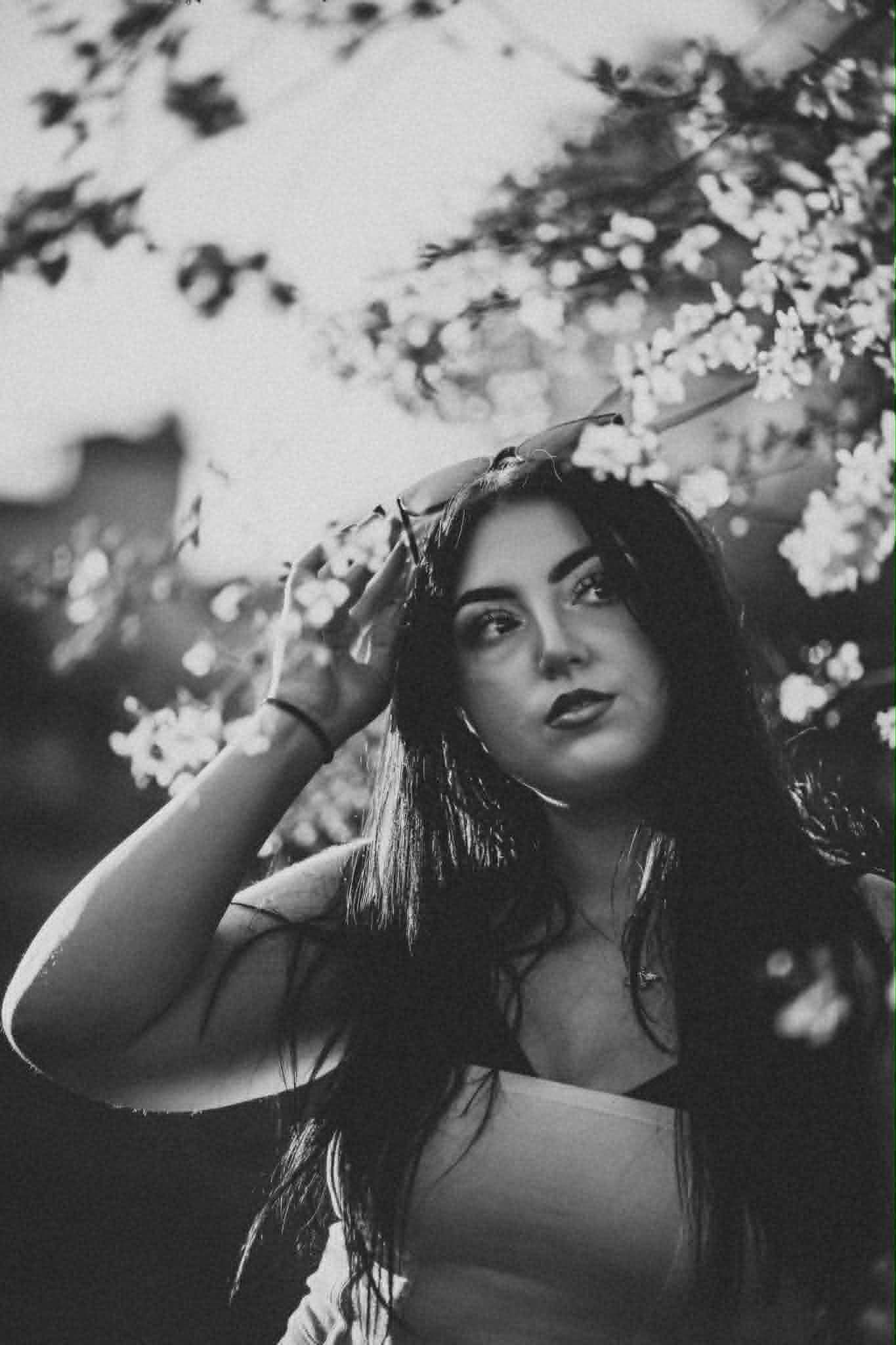 Black and white portrait of a young woman with long dark hair posing near blooming cherry blossom trees.