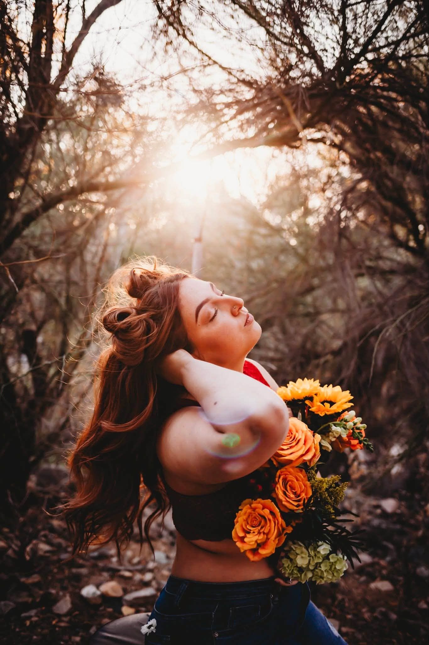 Woman with red hair holding orange roses and sunflowers, eyes closed, bathed in golden sunlight in a forest.