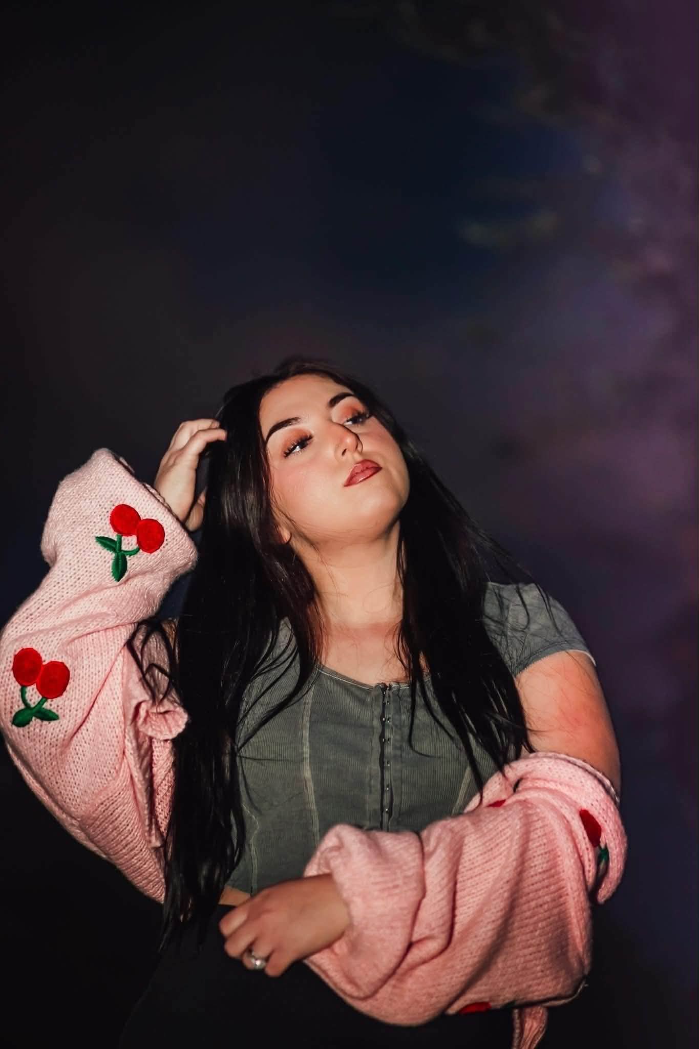 Young woman with long dark hair poses in a pink cherry-embroidered cardigan and gray top against a dark studio background.