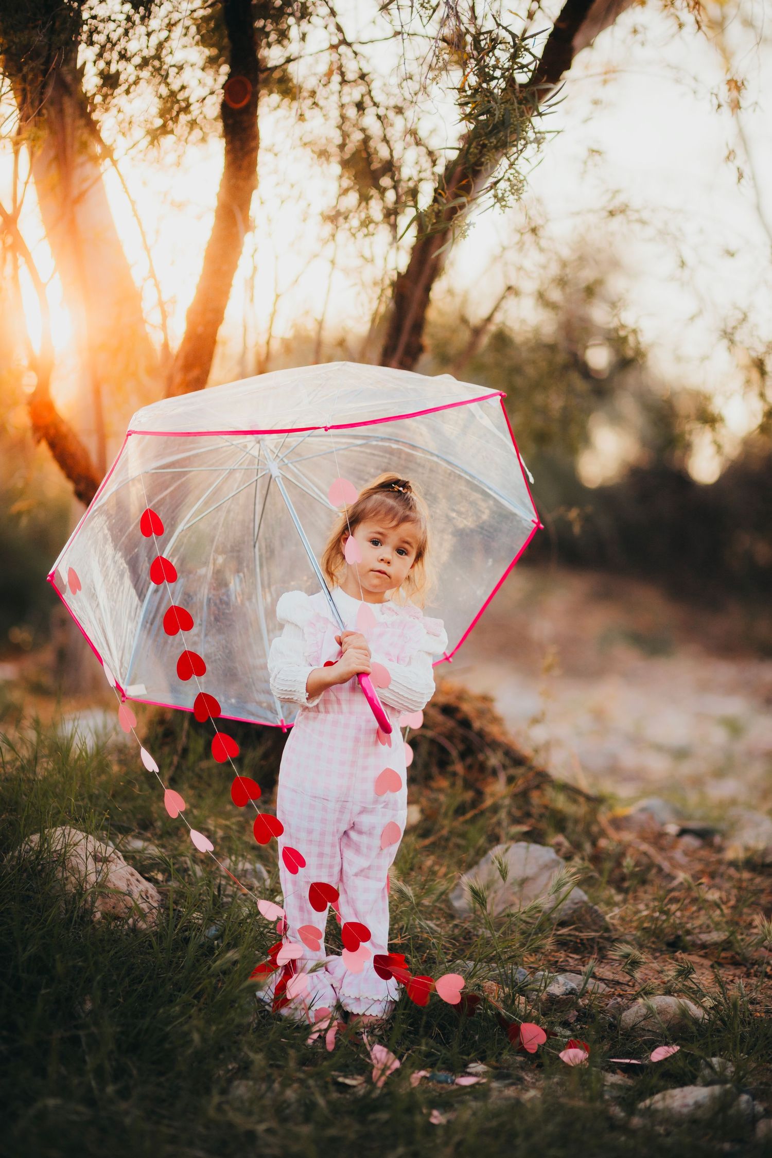 Young girl in pink outfit holds heart-decorated transparent umbrella outdoors at golden sunset.