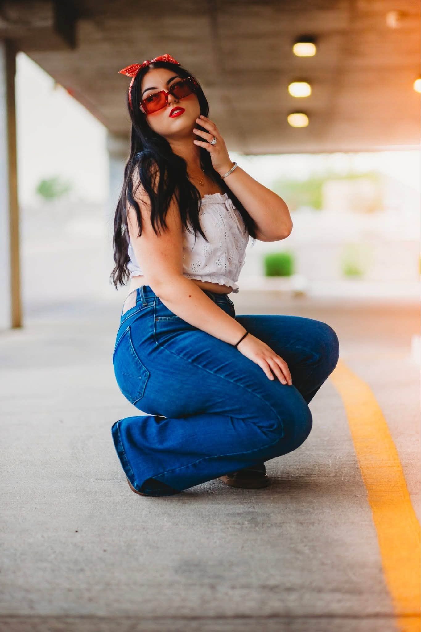 Young woman in vintage pin-up style, red sunglasses, white crop top, and wide-leg jeans, posing in a parking garage.
