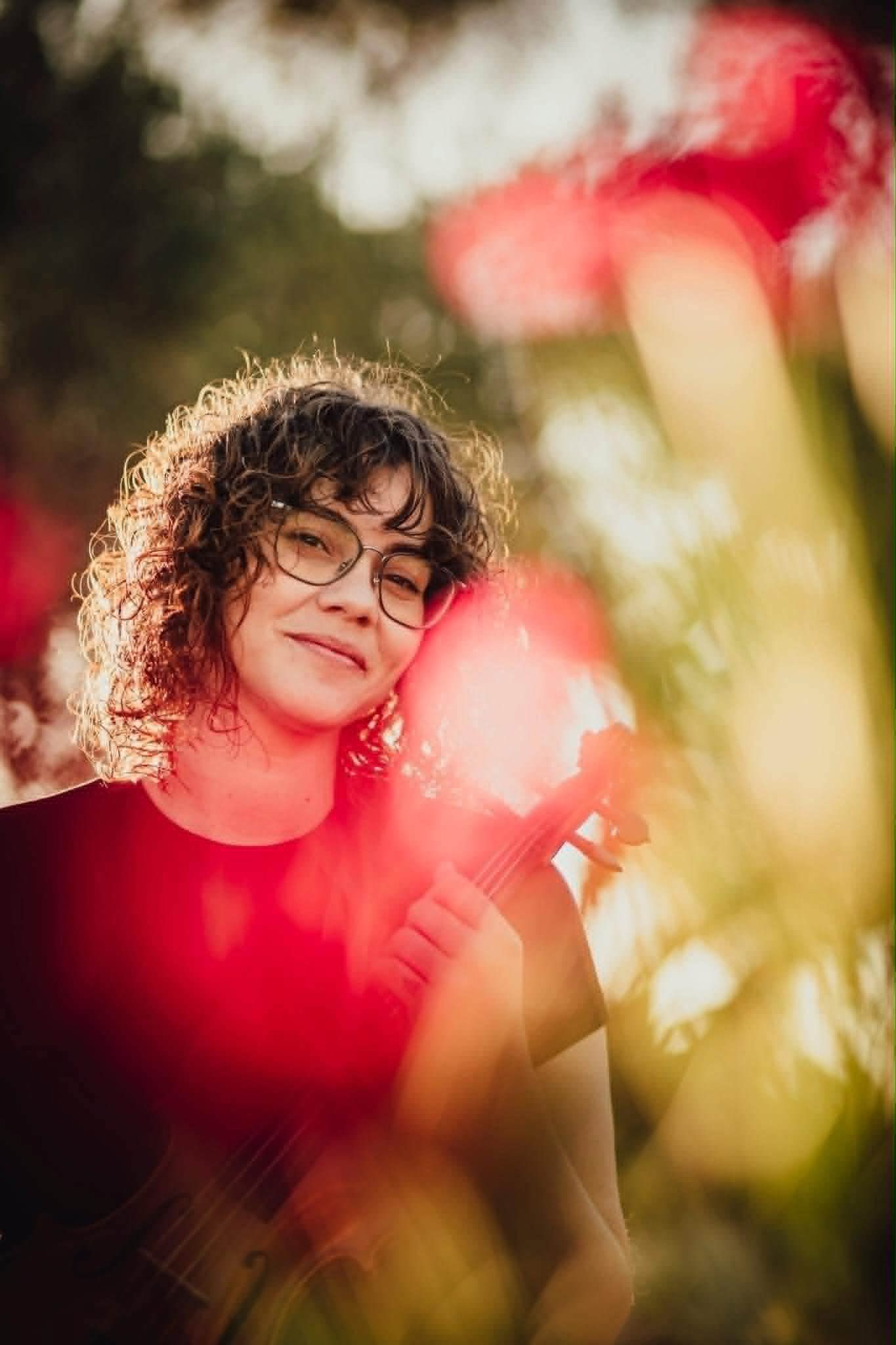 A curly-haired woman with glasses holds a violin outdoors, surrounded by soft red bokeh and warm golden sunlight.