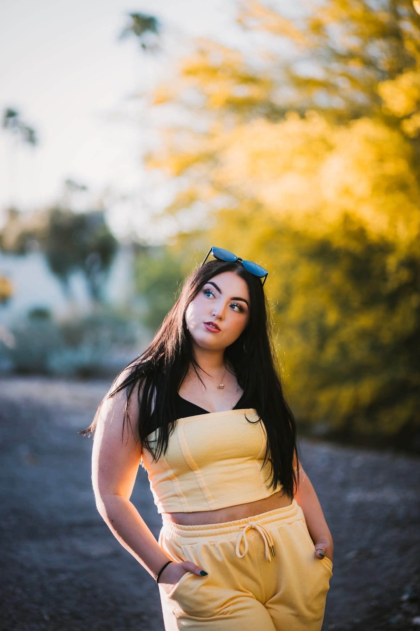 Young woman in yellow crop top and sweatpants with blue sunglasses poses outdoors among golden autumn trees.