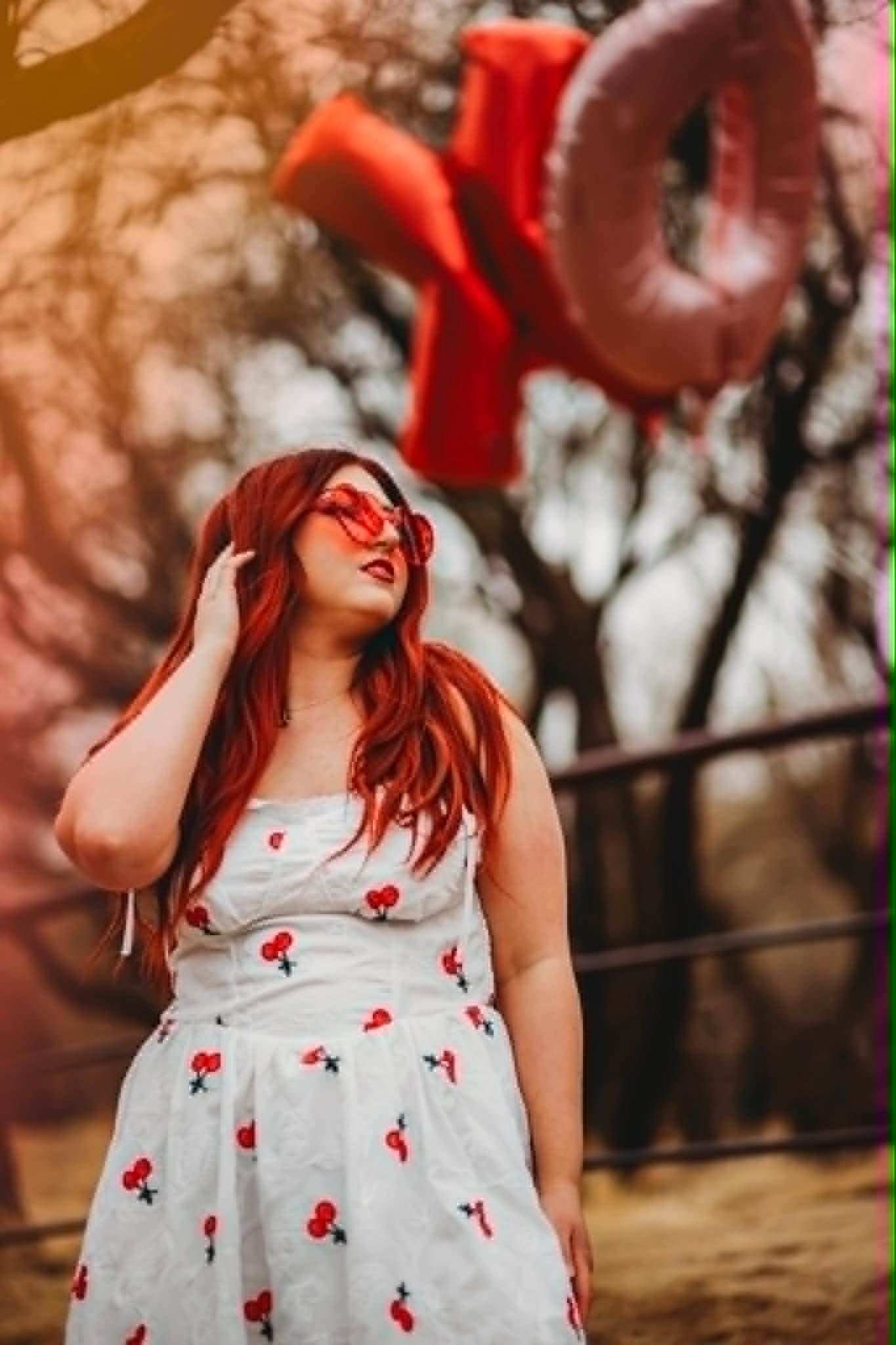 Woman with red hair in cherry print dress and red sunglasses poses near LOVE balloon letters outdoors.