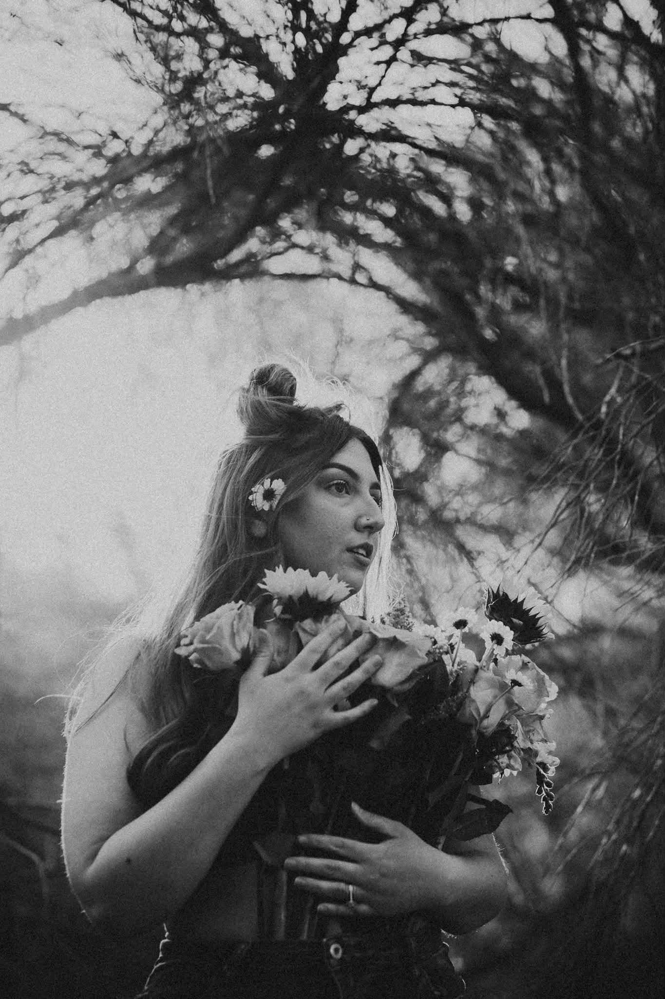 Black and white photo of a young woman holding wildflowers in her hair among bare trees in a dreamy forest setting.
