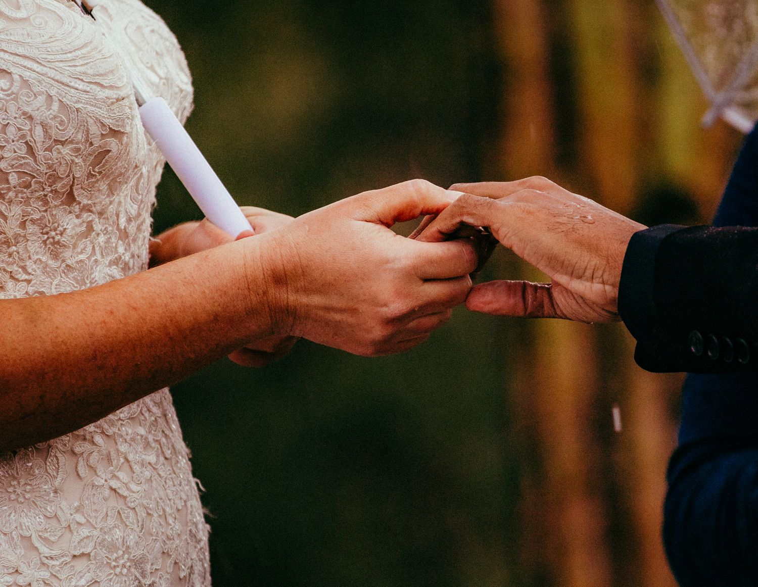 Bride and groom exchanging wedding rings during outdoor ceremony, close-up of hands placing ring on finger.