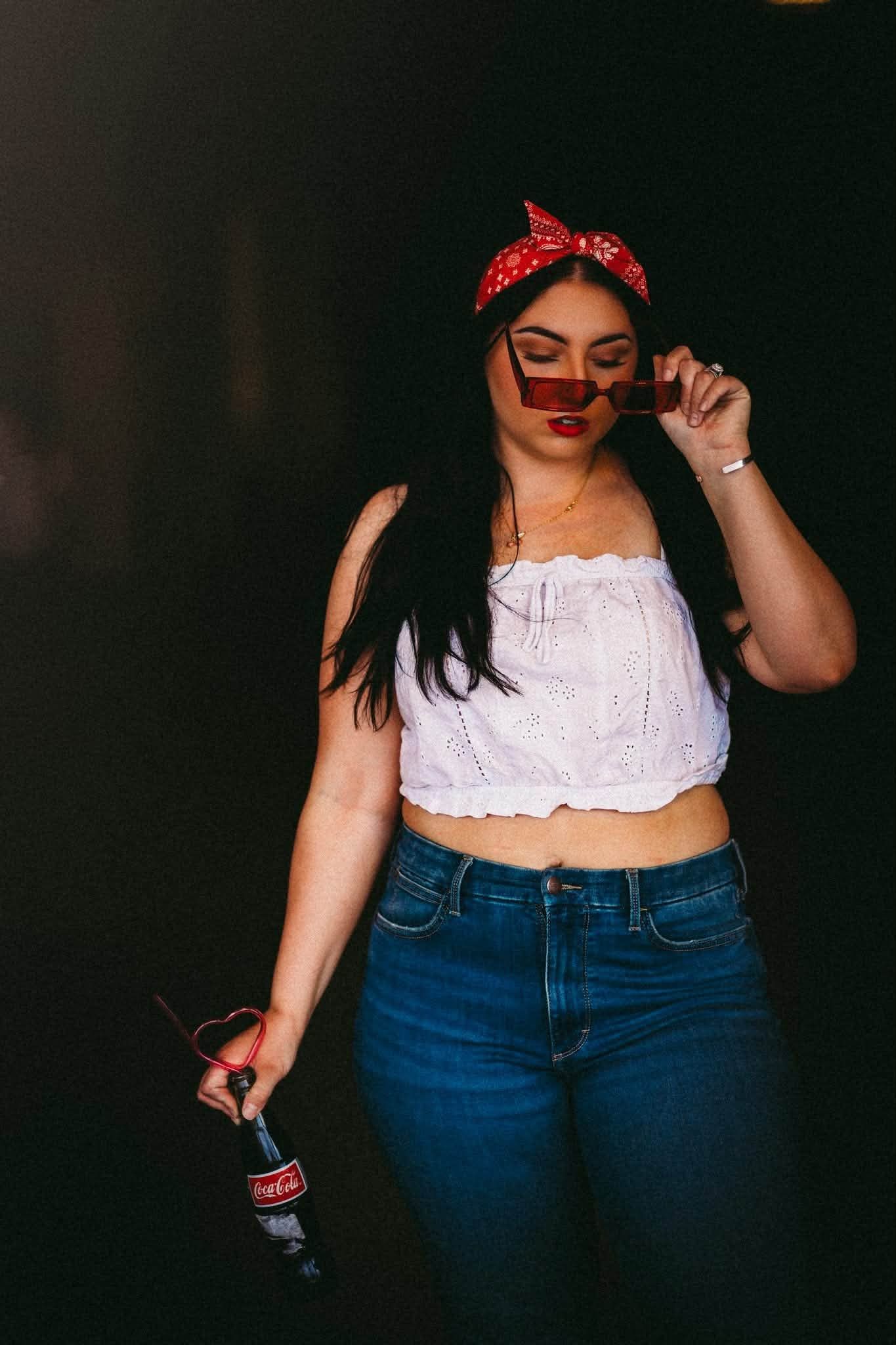 Young woman in white crop top, jeans, red bandana headband, and sunglasses holds a Coca-Cola bottle against dark background.