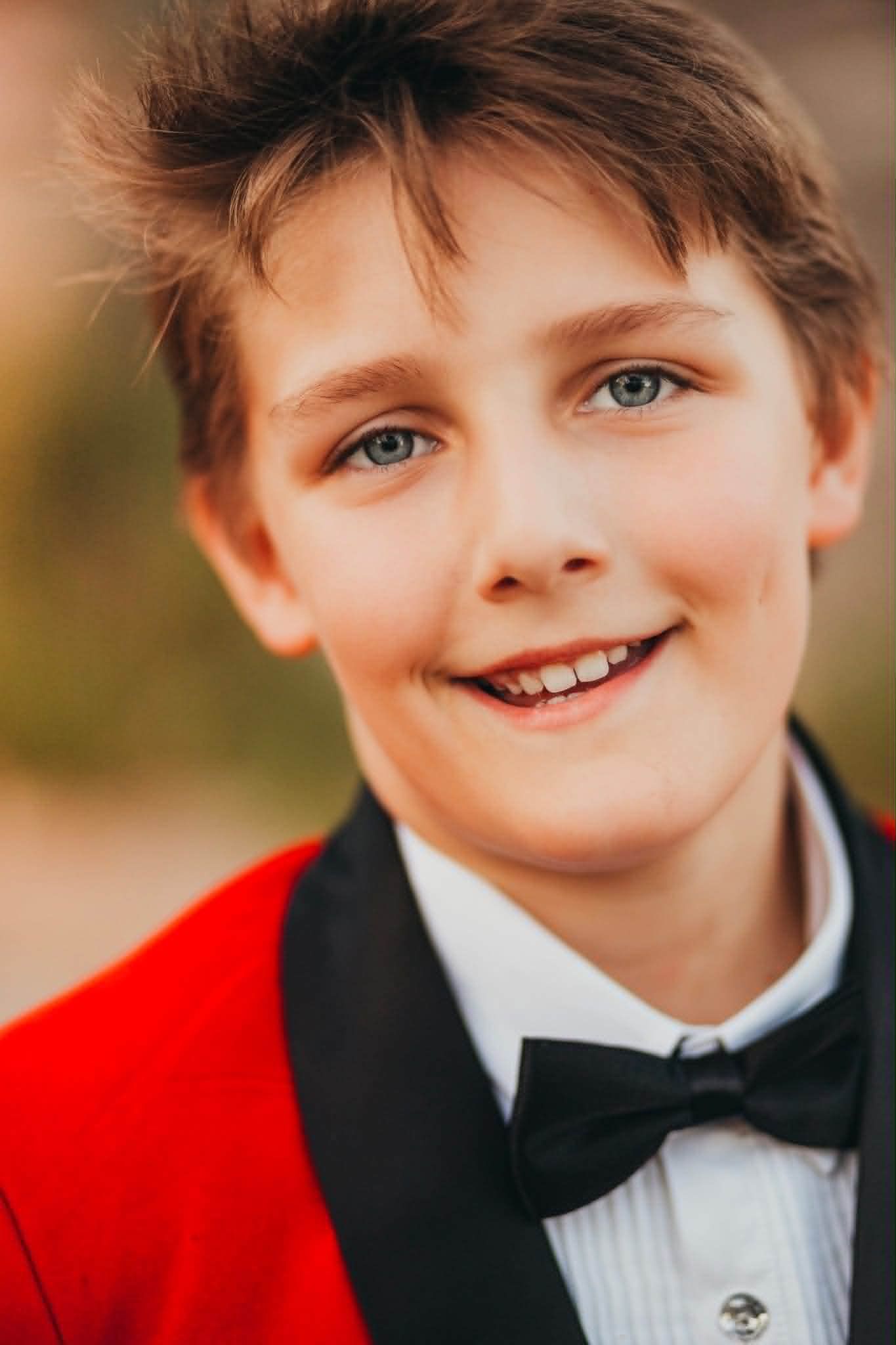 Smiling boy in a red tuxedo jacket with black bow tie, wearing a white dress shirt, portrait photo.