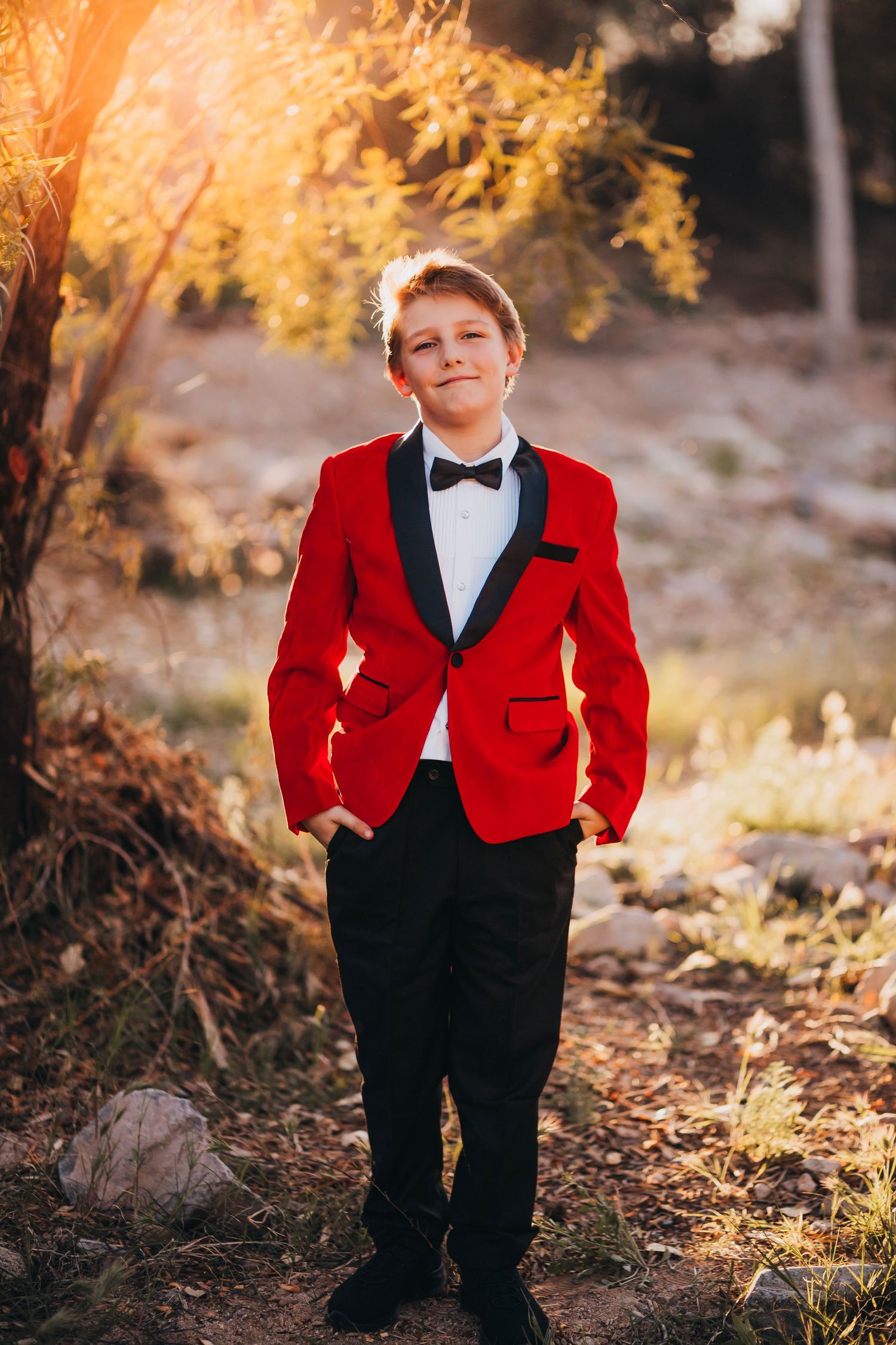 A young boy in a red tuxedo jacket with black lapels and bow tie poses outdoors in golden sunlight.