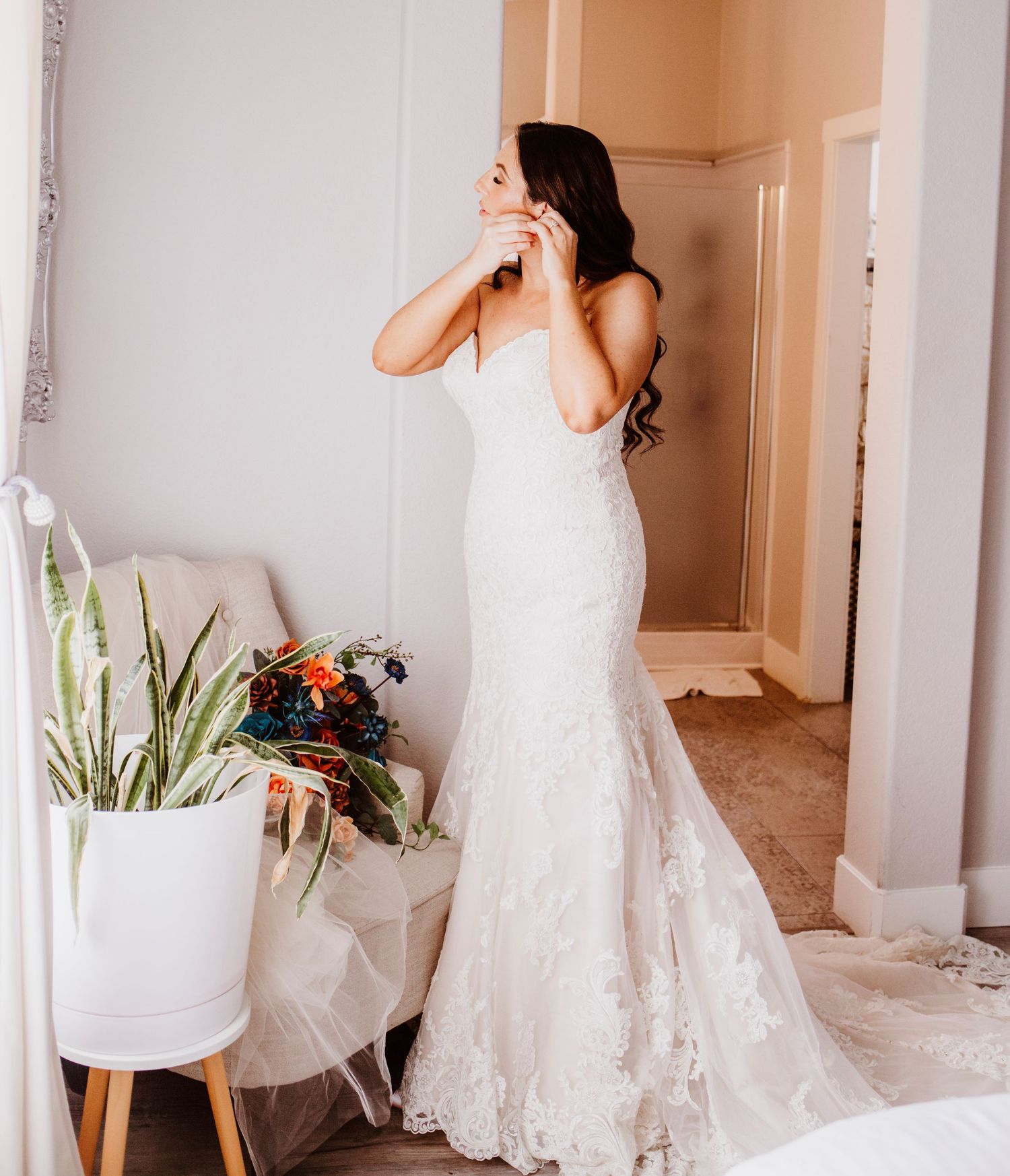 Bride in a strapless lace mermaid wedding gown with a train, posing in a bright room with a colorful bouquet nearby.