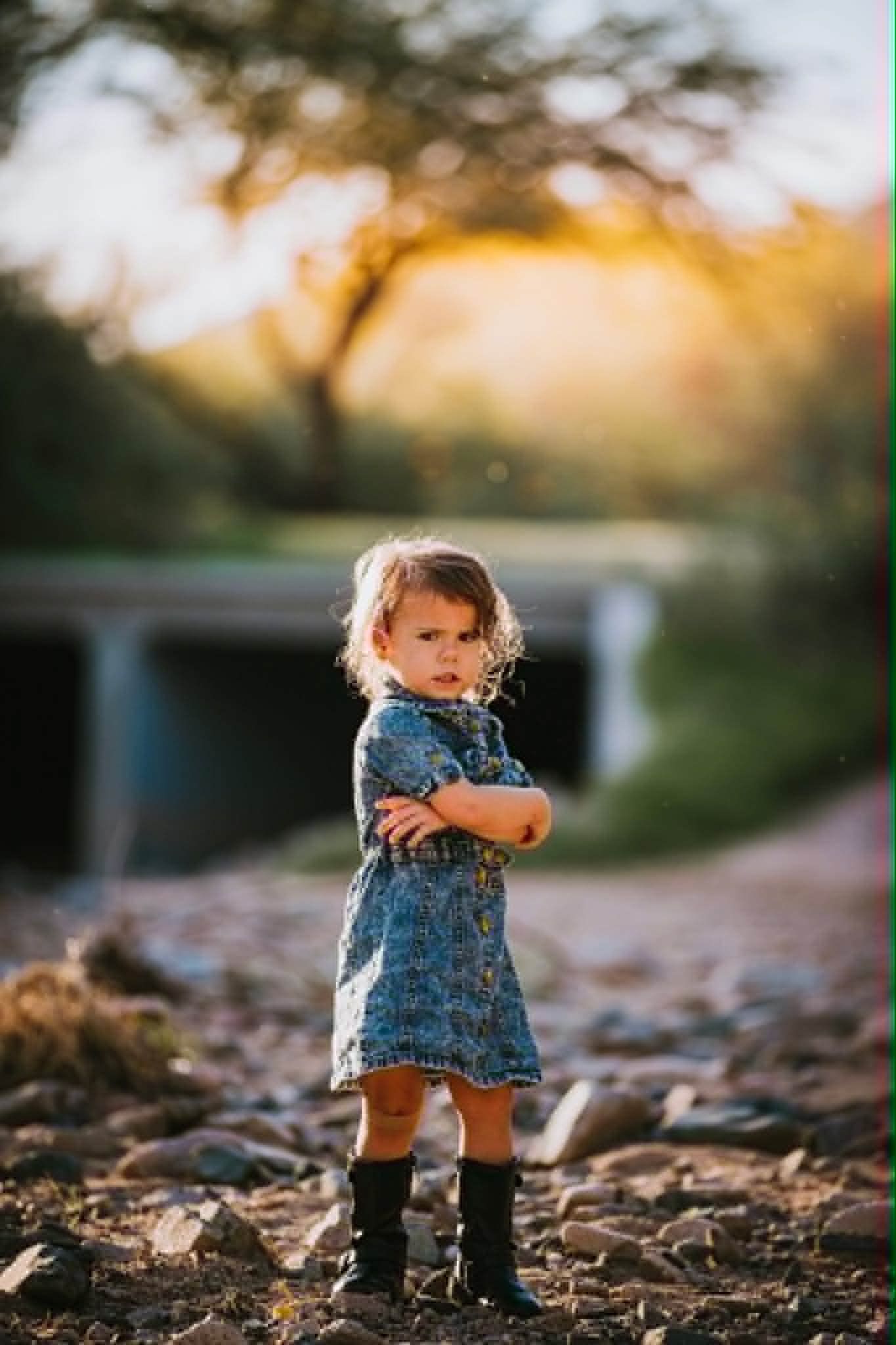 A young girl in a denim dress and black boots stands with arms crossed on a rocky path at golden hour.