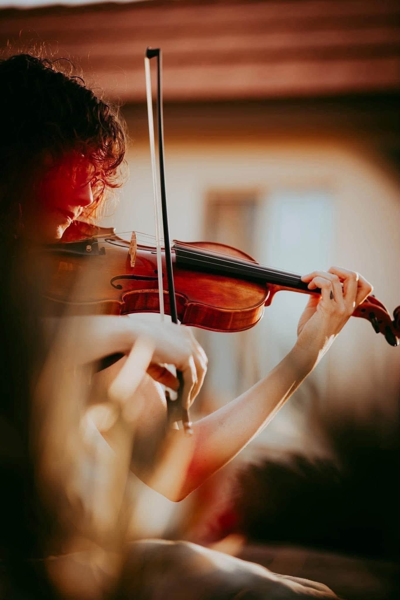A violinist playing a wooden violin with a bow, captured in warm, bokeh-lit close-up during a musical performance.
