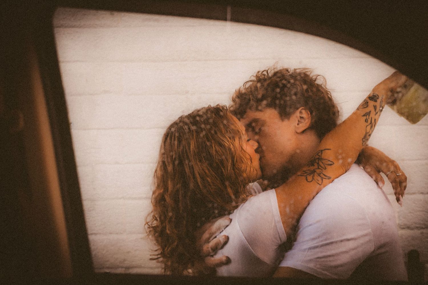 Couple sharing a romantic kiss inside a car, man with tattoos embracing curly-haired woman against white wall.