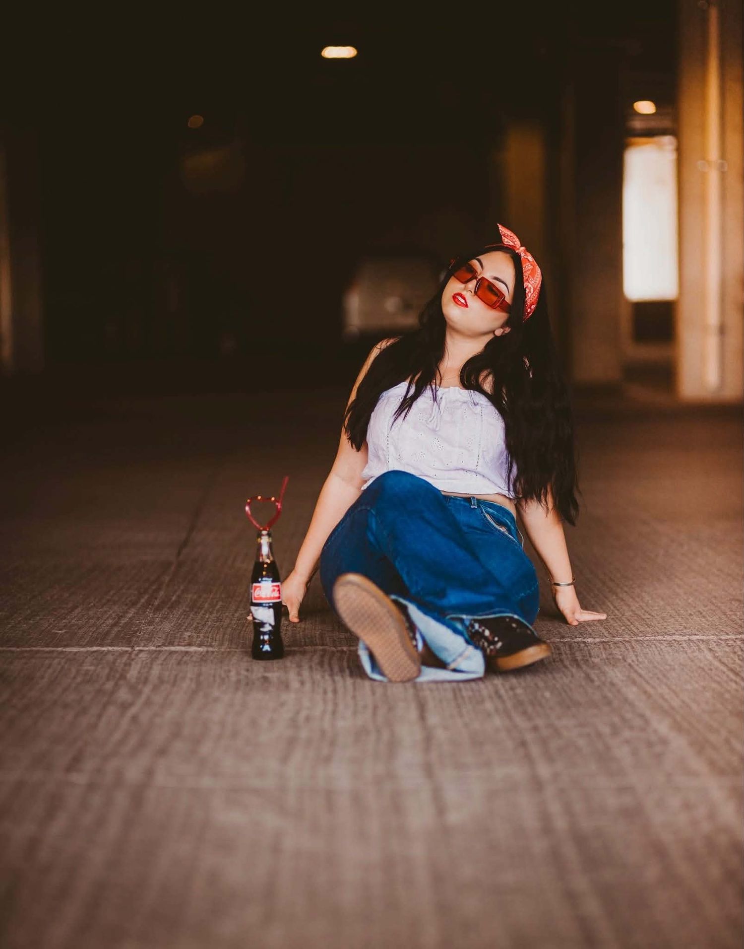 Young woman in denim skirt and red sunglasses sitting on ground with a bottle in moody urban setting.