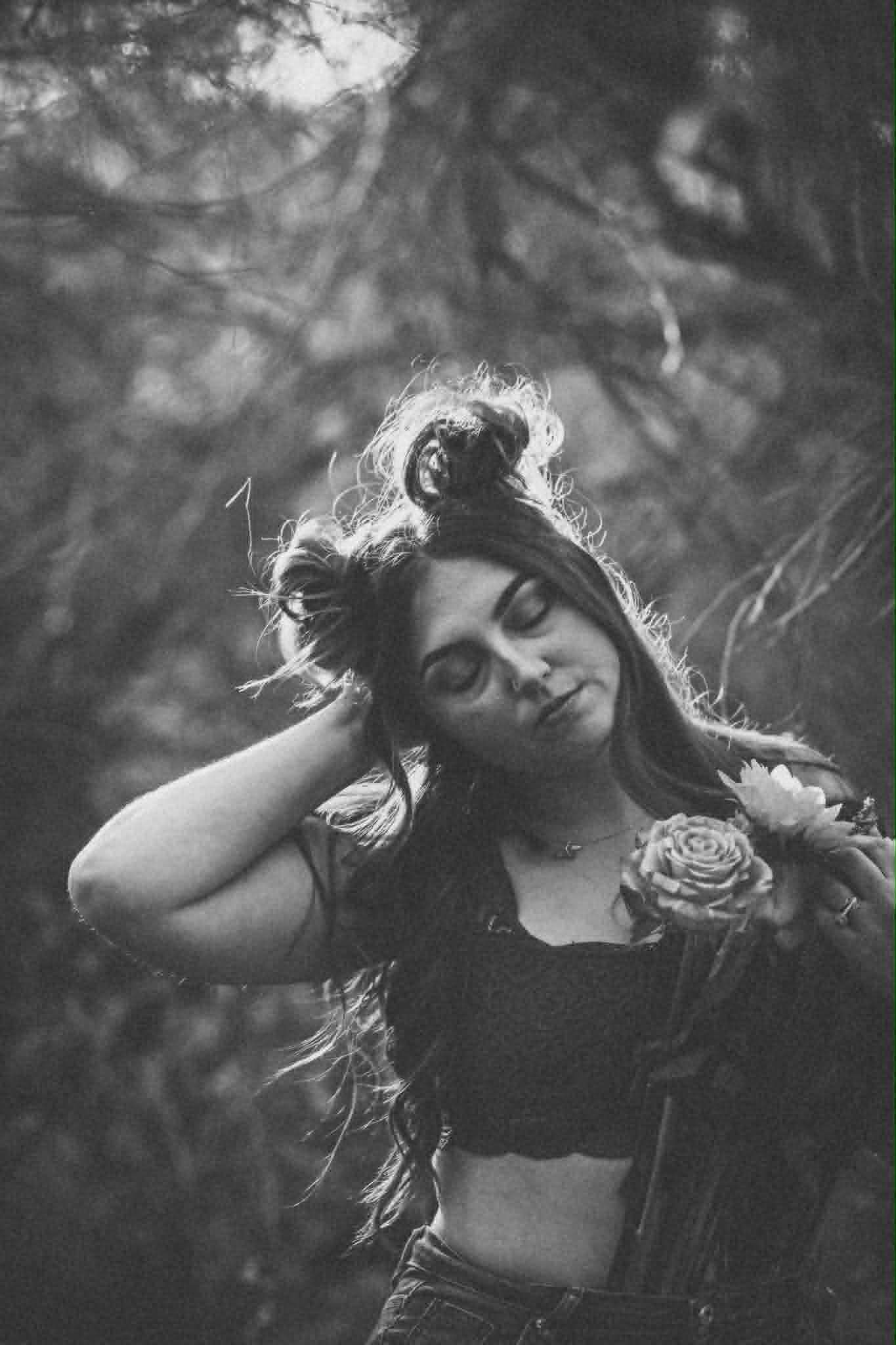 Black and white portrait of a young woman with flowing hair holding a rose, eyes closed, surrounded by trees.