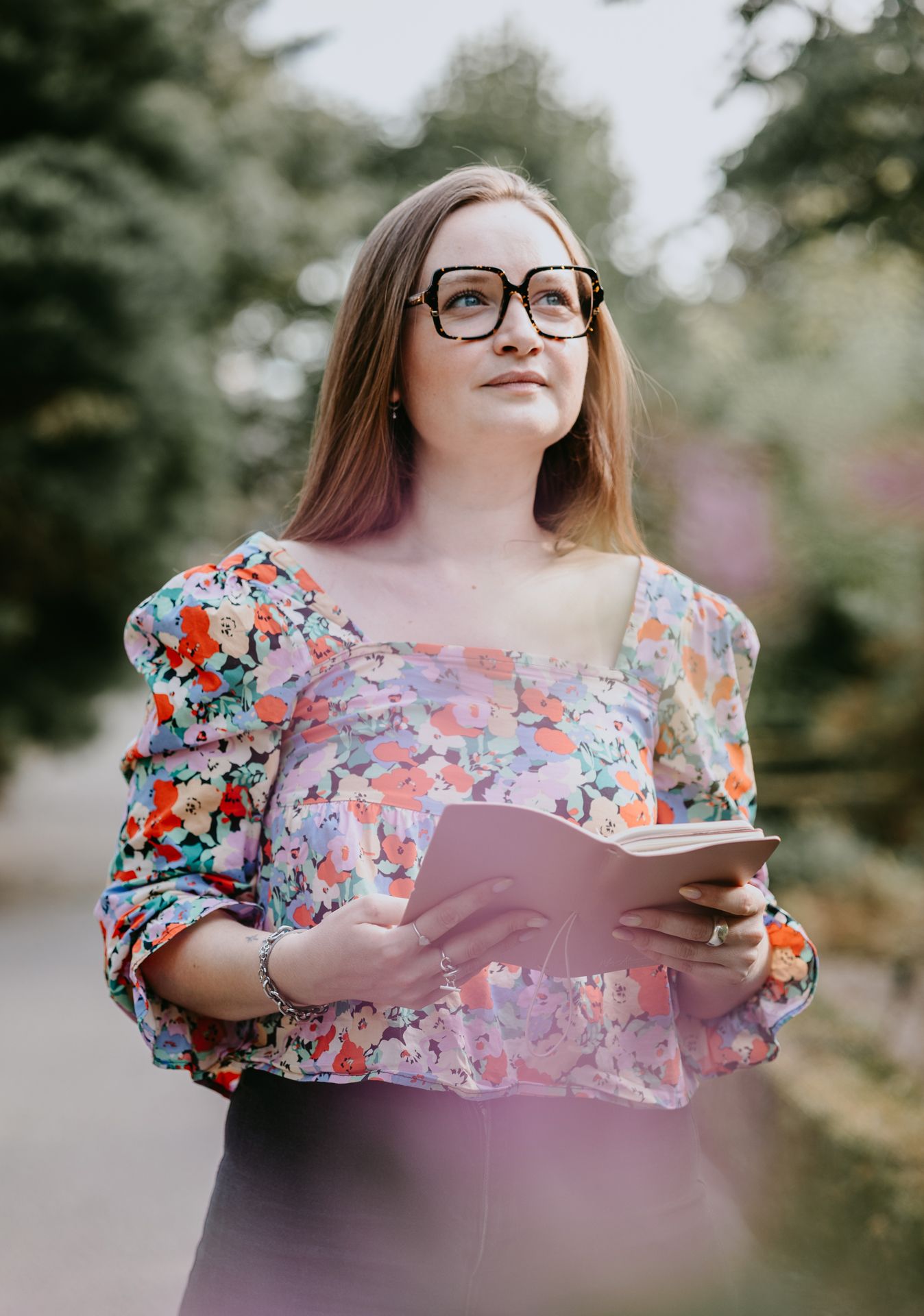 Young woman in floral blouse and glasses holding an open book outdoors in a garden setting.
