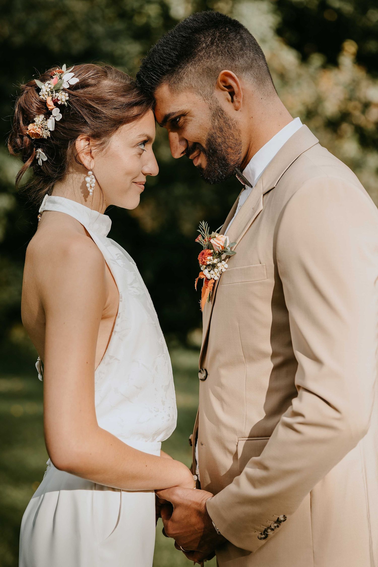 Smiling bride and groom in beige suit touch foreheads tenderly in outdoor garden setting.