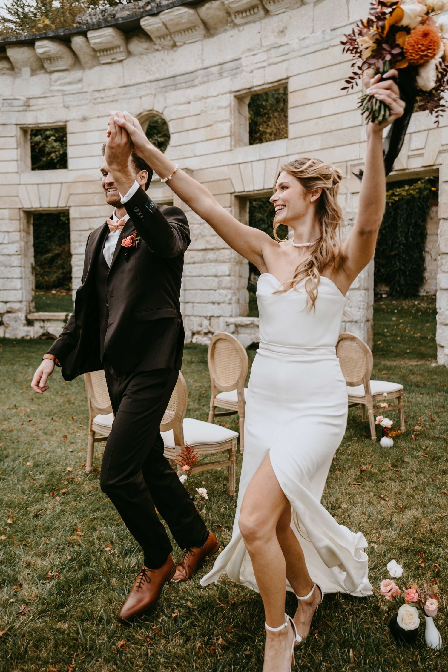 Joyful newlyweds celebrate after outdoor wedding ceremony, bride in white gown raises bouquet while groom spins her.