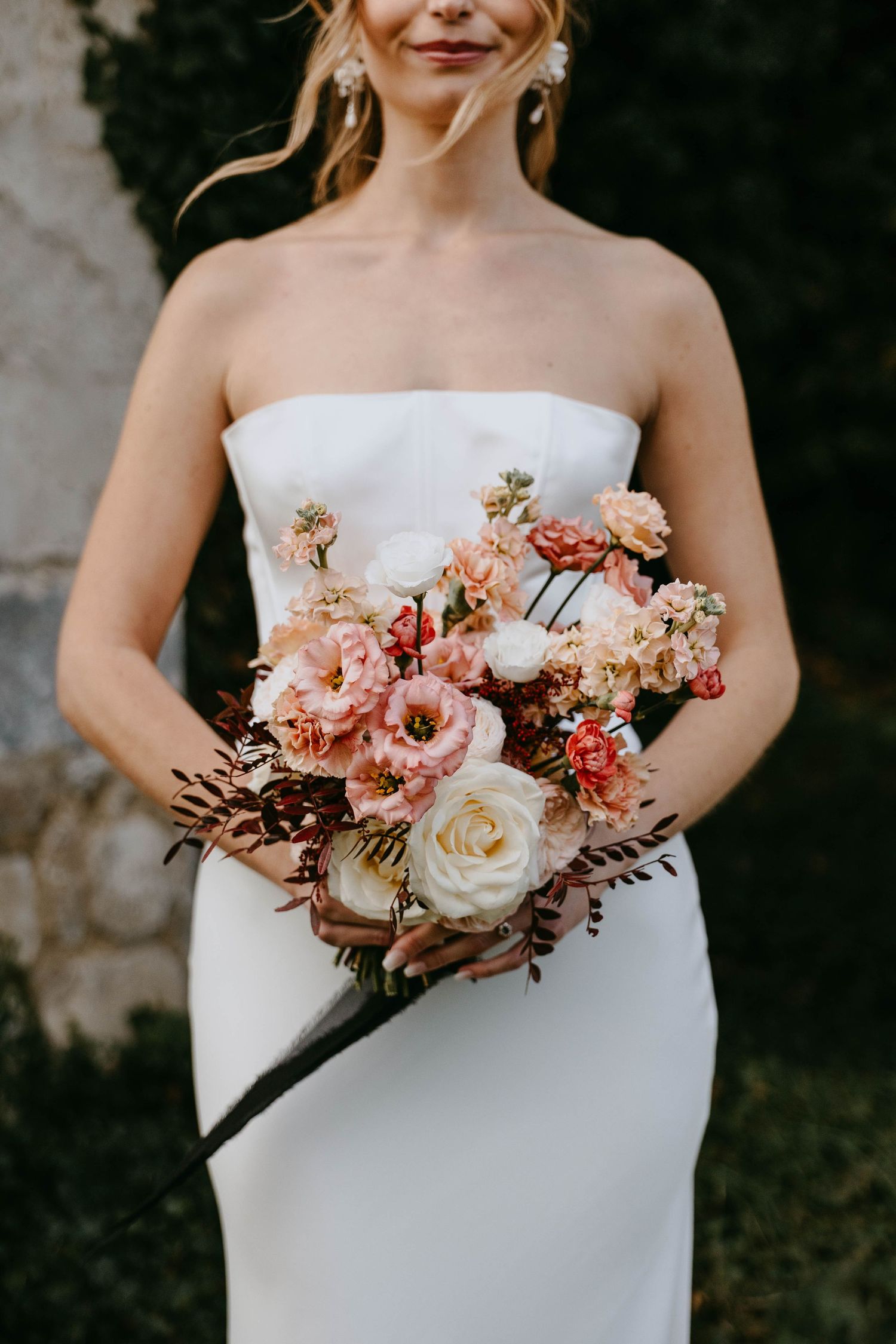Bride in strapless white gown holding lush bridal bouquet with blush pink, cream roses, and dark foliage.