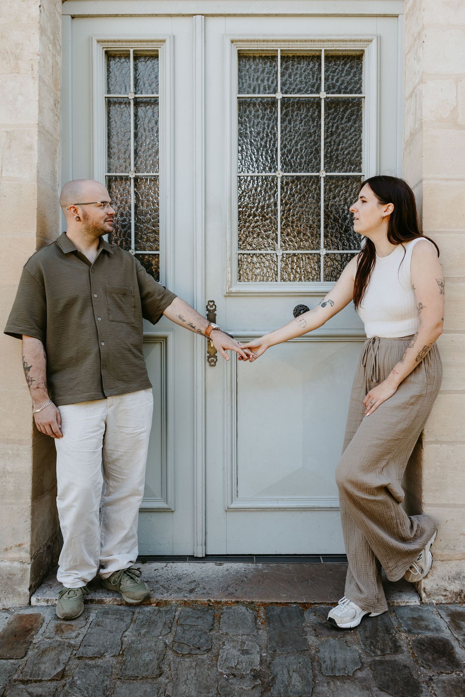 Couple holding hands in front of a sage green door on cobblestone, man in olive shirt, woman in white top and tan pants.