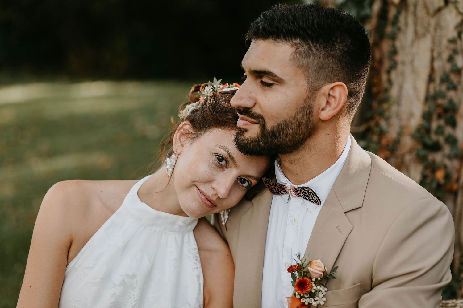 Romantic wedding couple posing outdoors, bride in white dress resting head on groom's shoulder in a tender embrace.