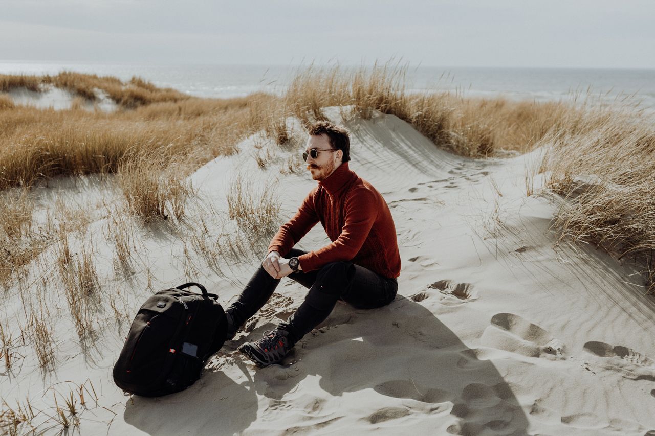Man in red sweater sitting on sandy beach dunes with a backpack beside him on a cloudy day.
