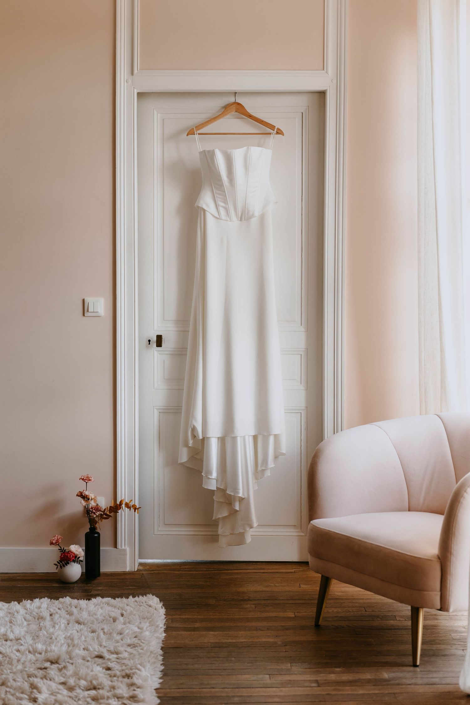 Elegant white strapless wedding dress hanging on wooden hanger against cream door beside blush pink velvet accent chair.