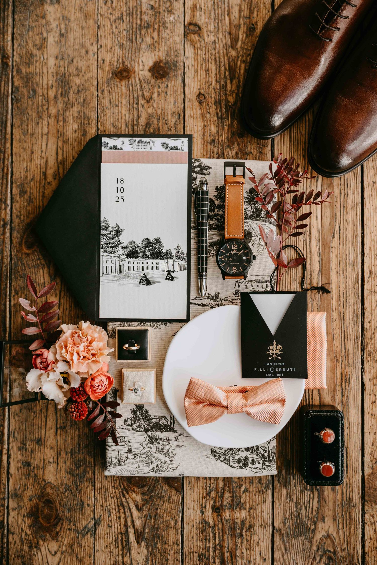 Elegant wedding flat lay with bow tie, watch, cufflinks, invitation suite, floral arrangement, and brown leather shoes on rustic wood.