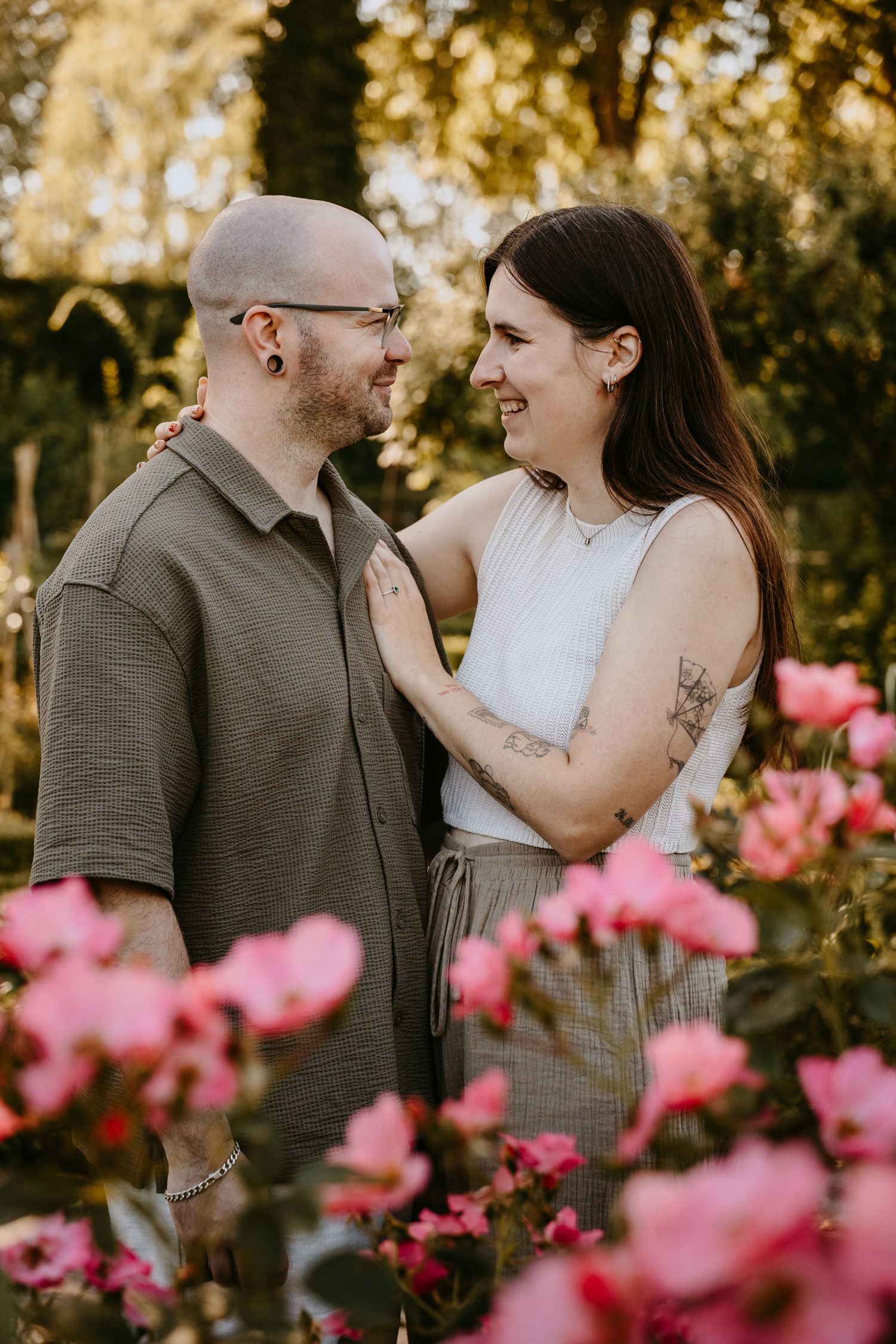 Couple smiling at each other in a garden surrounded by pink roses, woman with tattoos resting hand on mans chest.