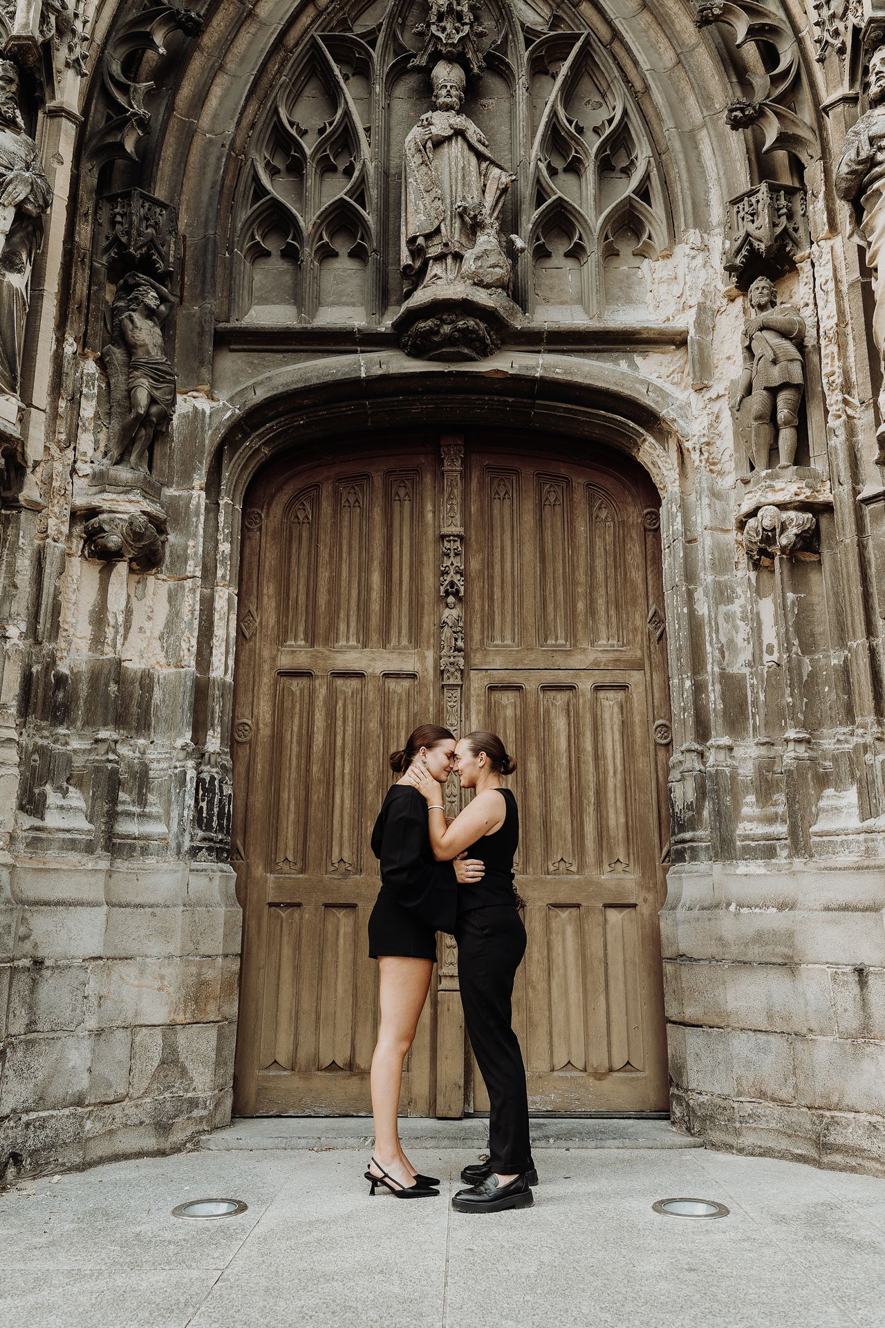 Couple sharing a kiss in front of an ornate Gothic cathedral entrance with large wooden doors.