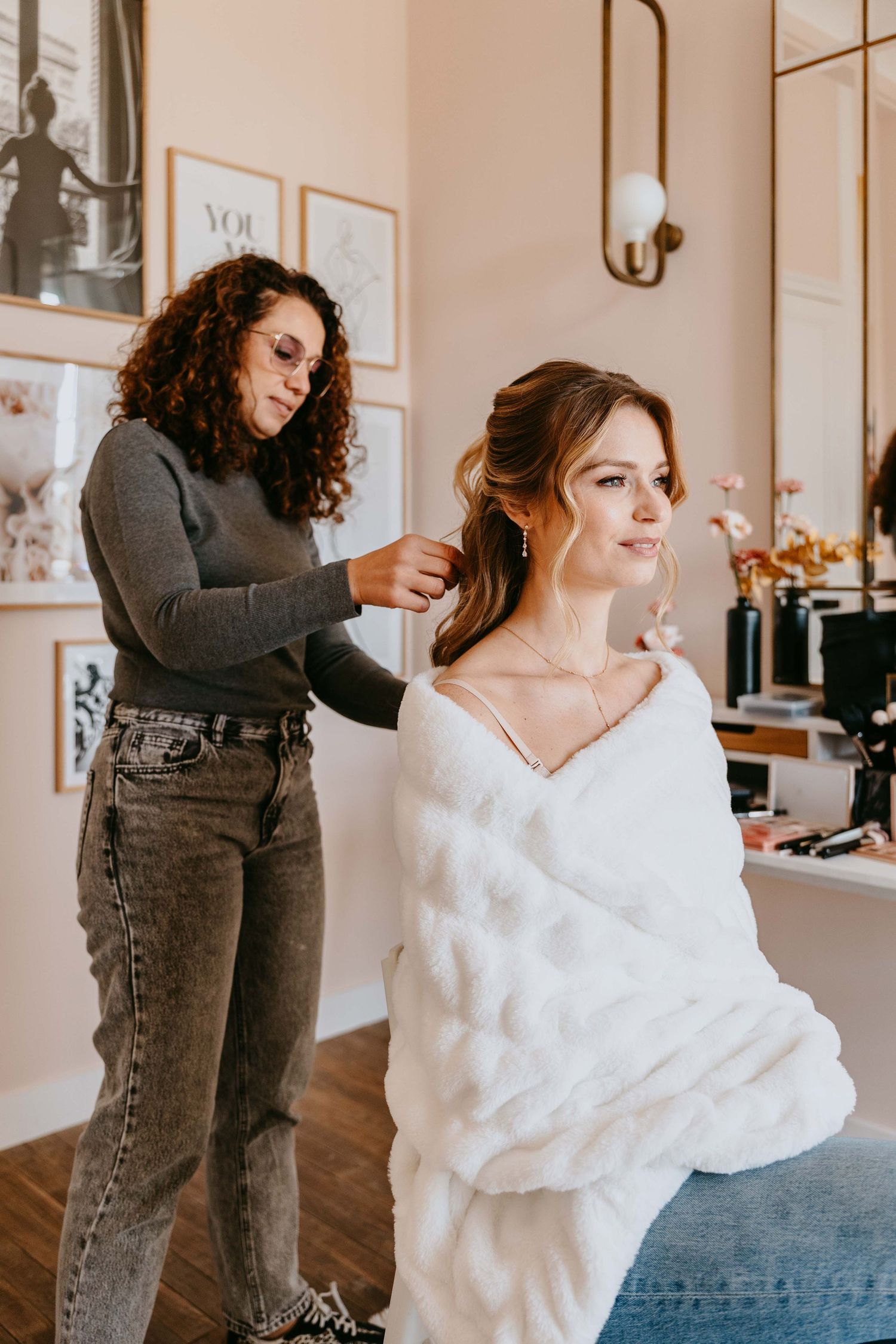 Professional hair stylist styling elegant bride in white fur shawl at a beautifully decorated beauty salon.