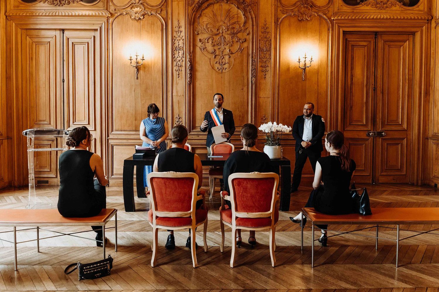 Wedding civil ceremony in an ornate wood-paneled room with guests seated before an officiant.
