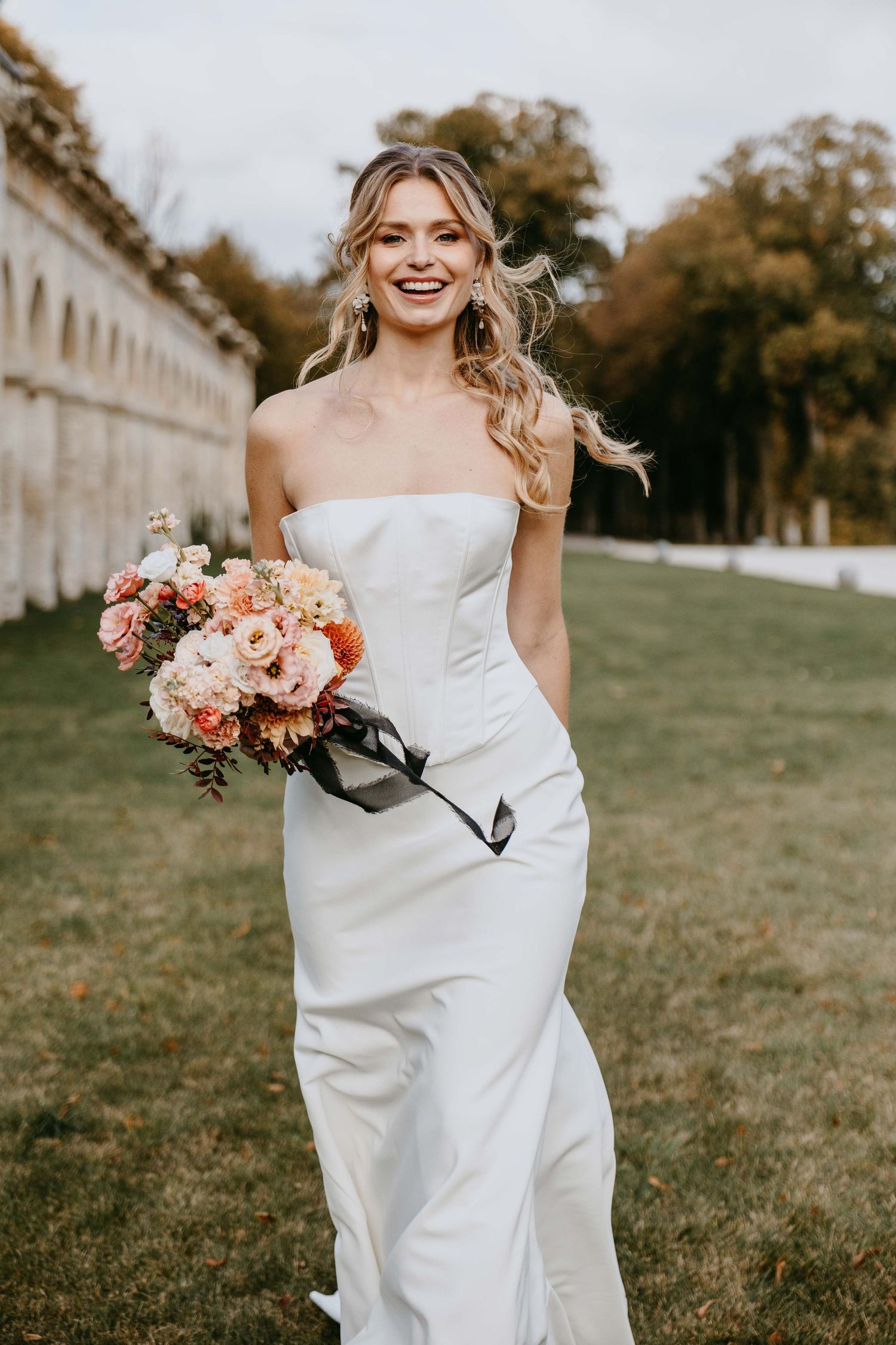 Smiling bride in strapless white gown holding peach and pink floral bouquet outdoors near stone architecture.