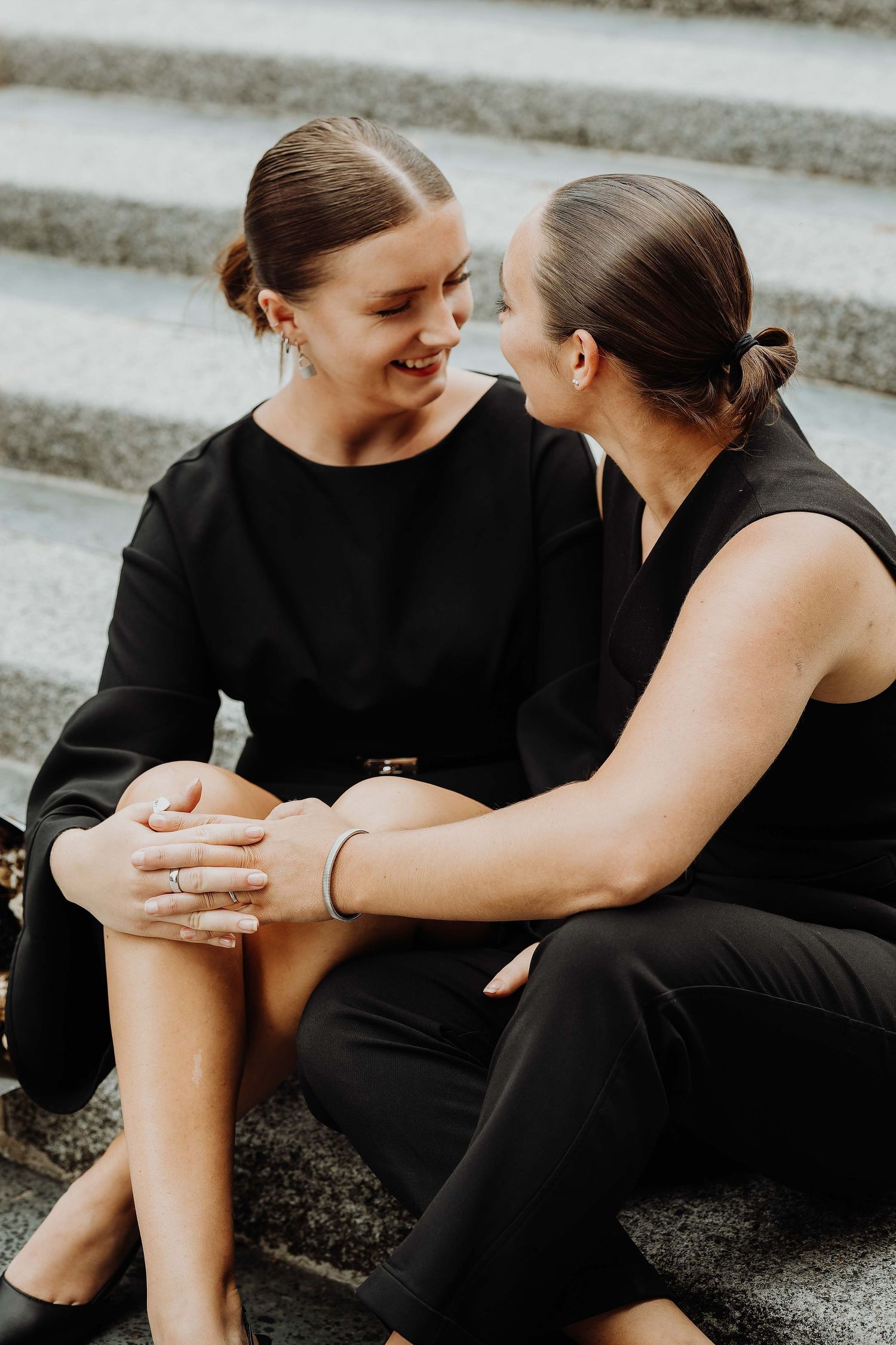 Two women in matching black outfits sitting on steps, smiling and sharing an intimate moment together.