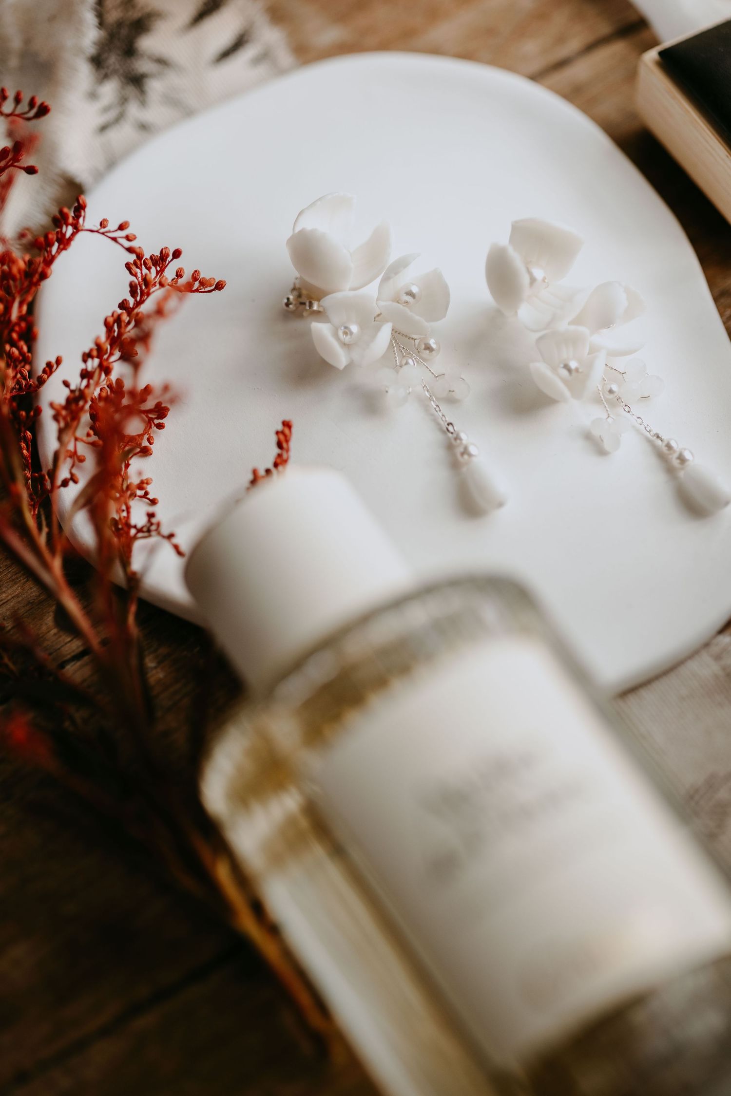 White floral earrings with crystal drops displayed on a ceramic plate beside red dried flowers and a glass bottle.