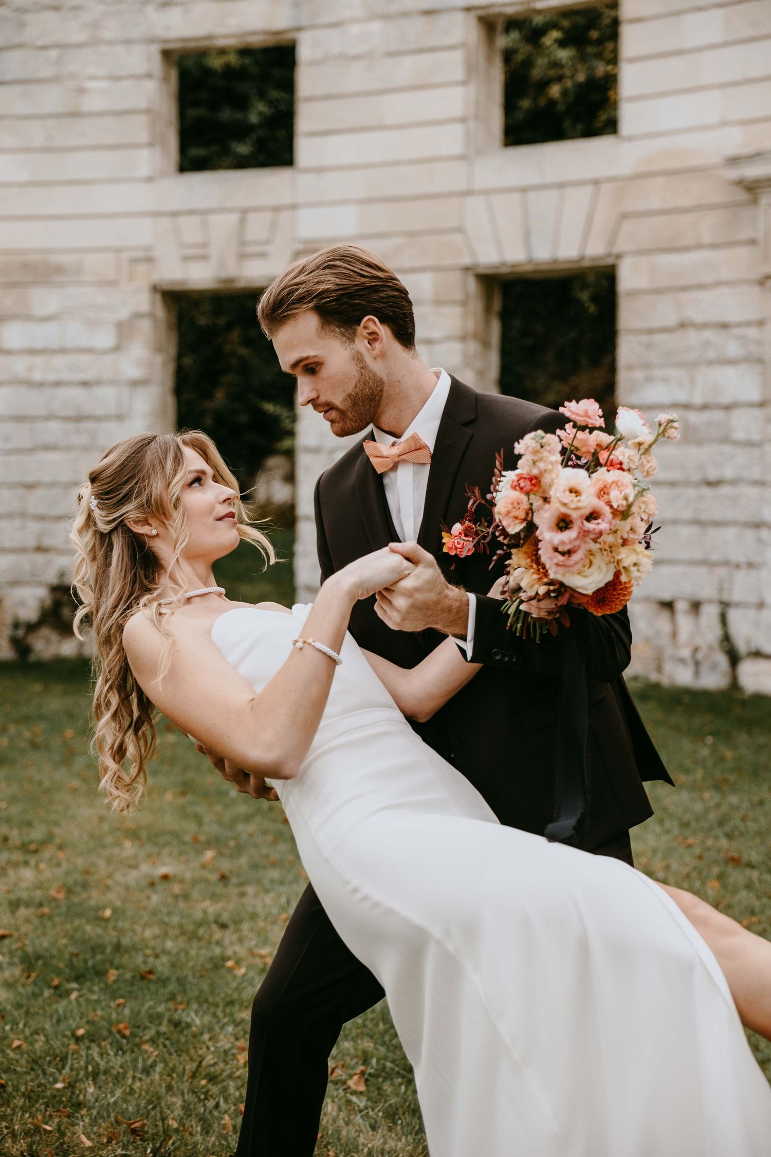 Groom in black tuxedo dips bride in white dress holding coral bouquet outdoors near stone building.