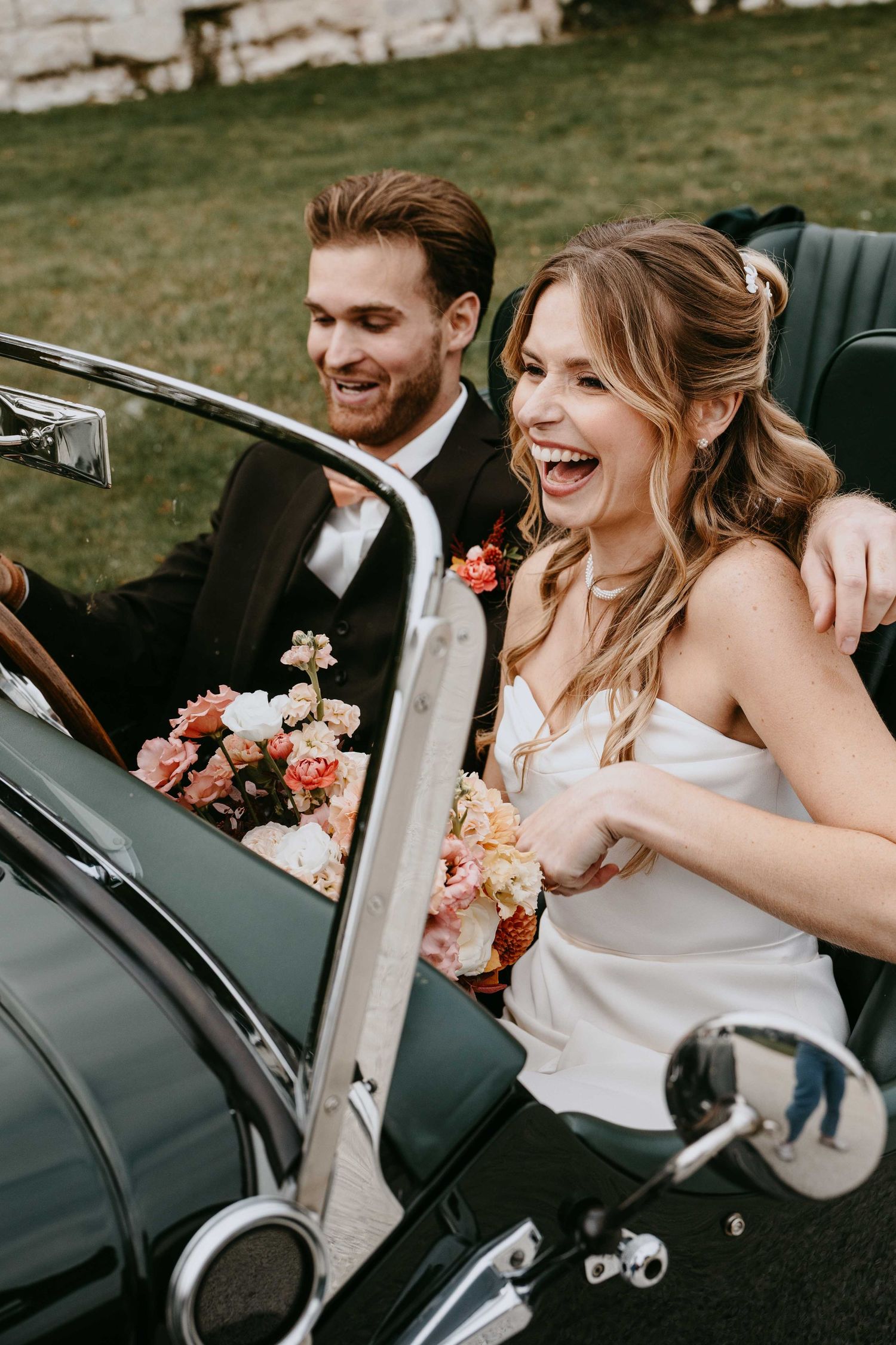 Joyful bride and groom laughing in a vintage green convertible car, bride holding a pink floral bouquet on their wedding day.