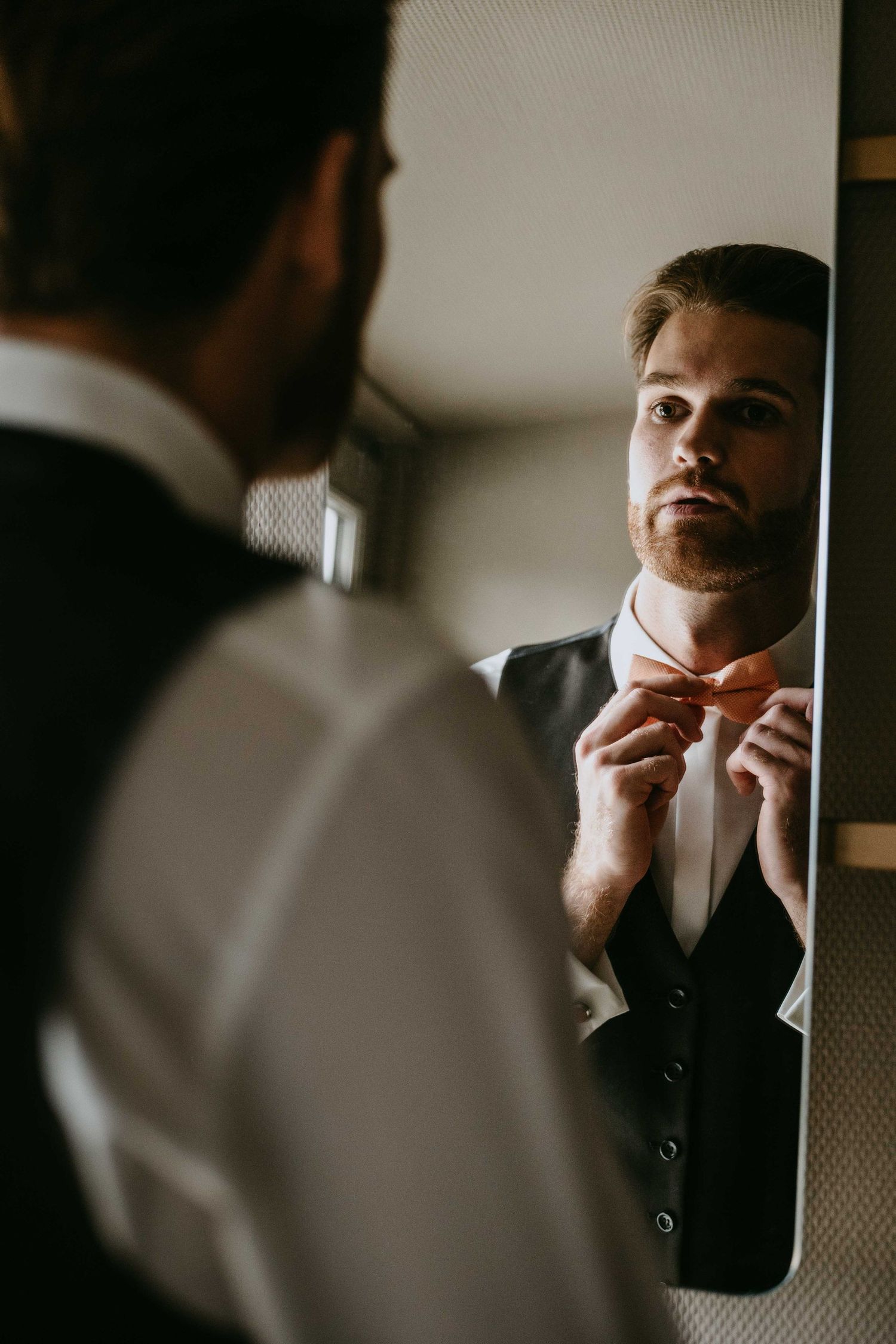 A bearded groom adjusts his bow tie while gazing at his reflection in a mirror, dressed in a white shirt and vest.