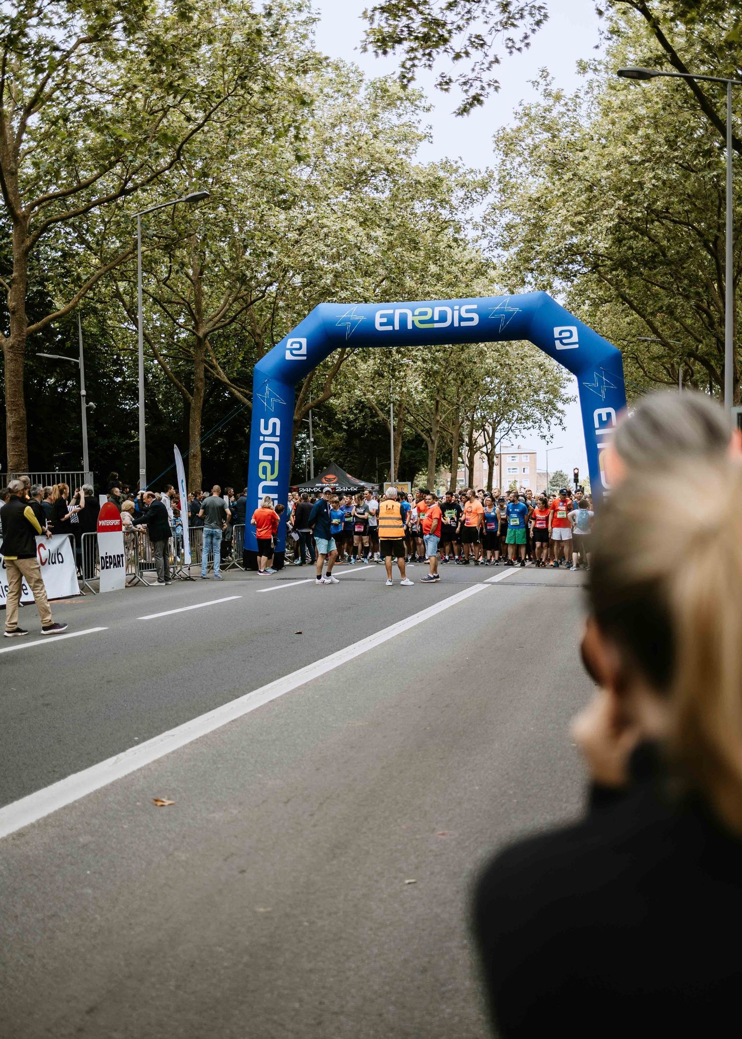 Runners gather at the start line of a road race under a blue Enedis inflatable arch surrounded by trees.
