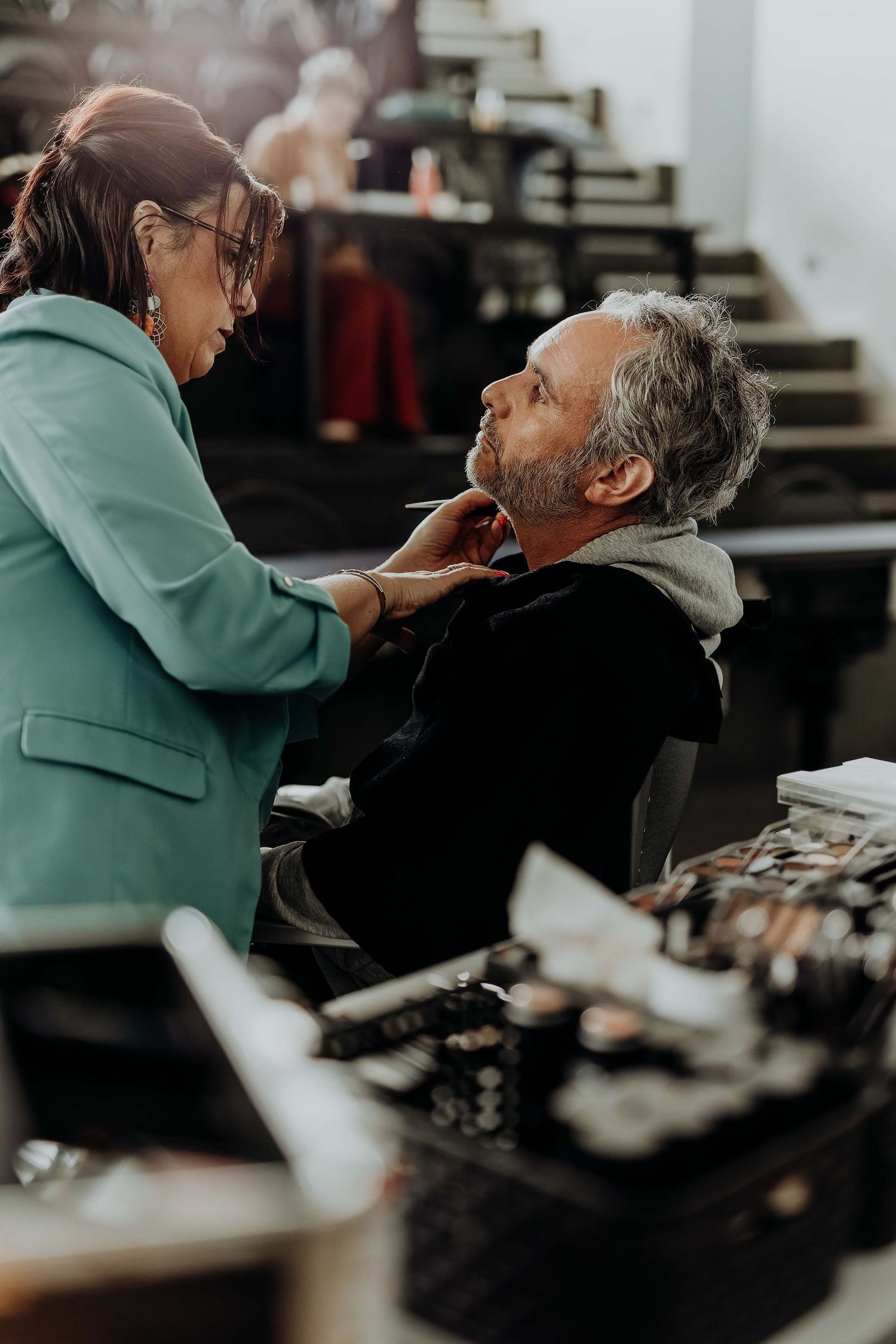 Makeup artist in teal blazer applying cosmetics to a seated man in a professional backstage setting.