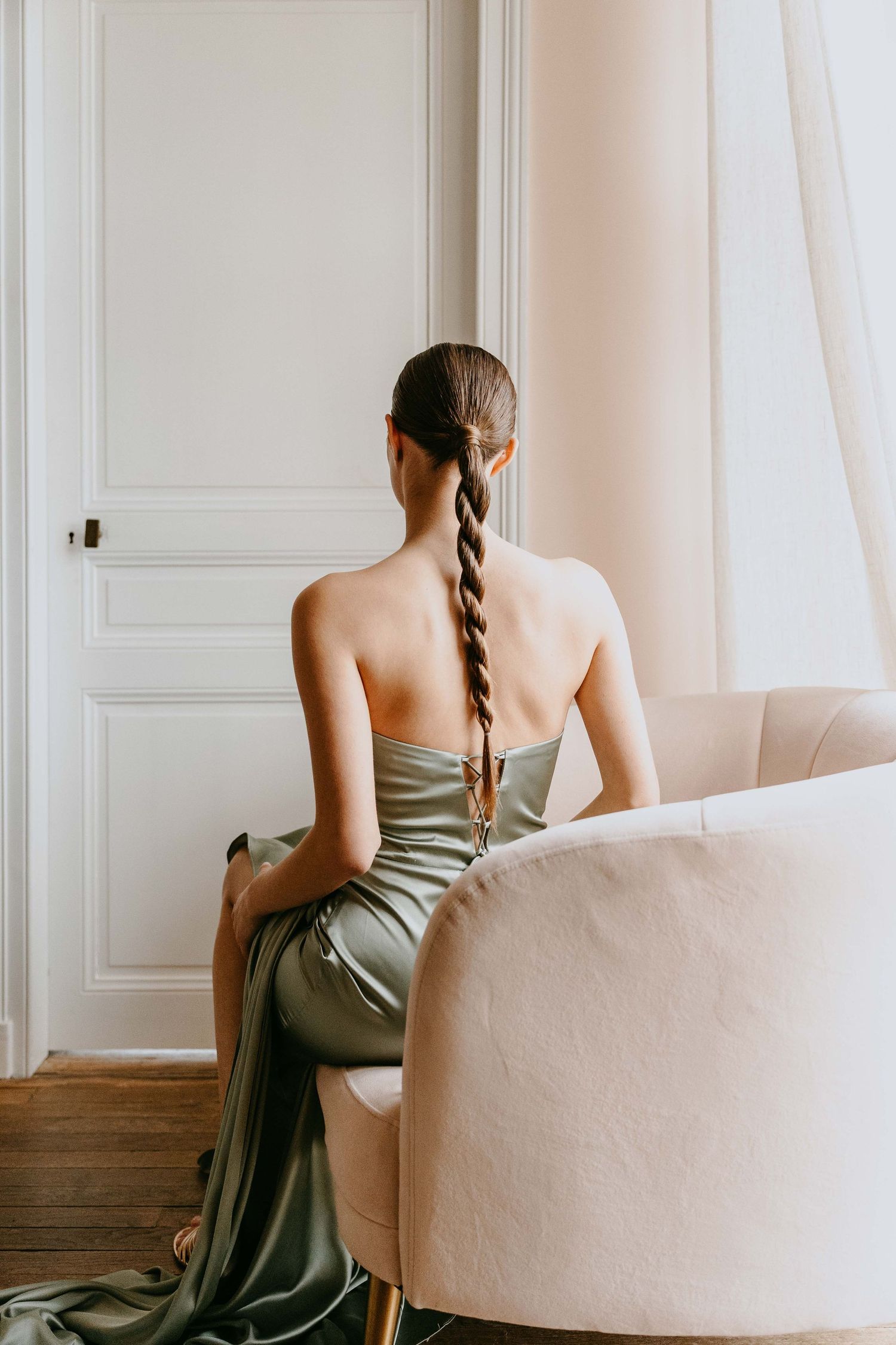 Woman in elegant olive satin strapless gown with long braid, seated in cream armchair facing white paneled wall.