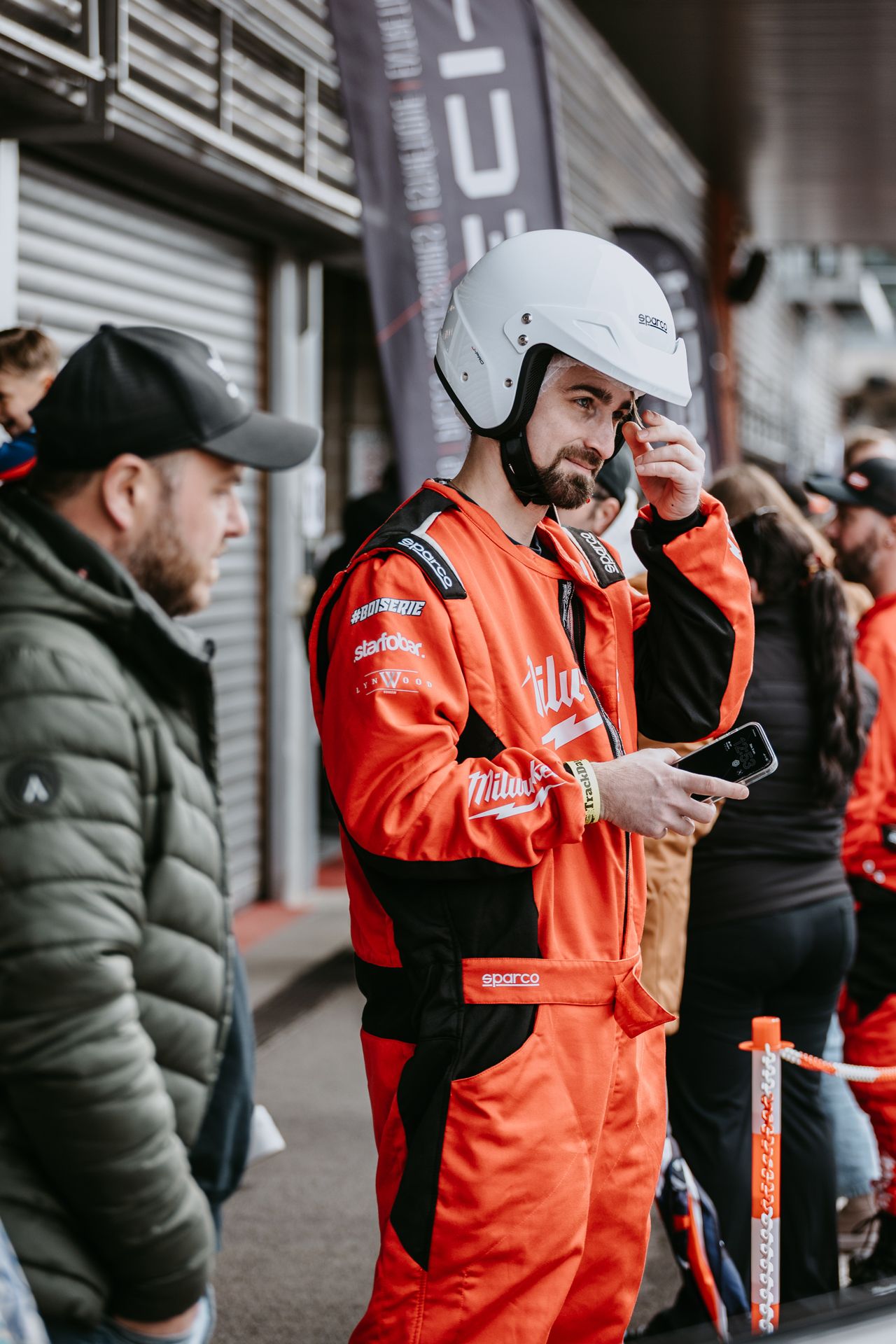 Bearded man in orange racing suit adjusting white helmet at motorsport event paddock.