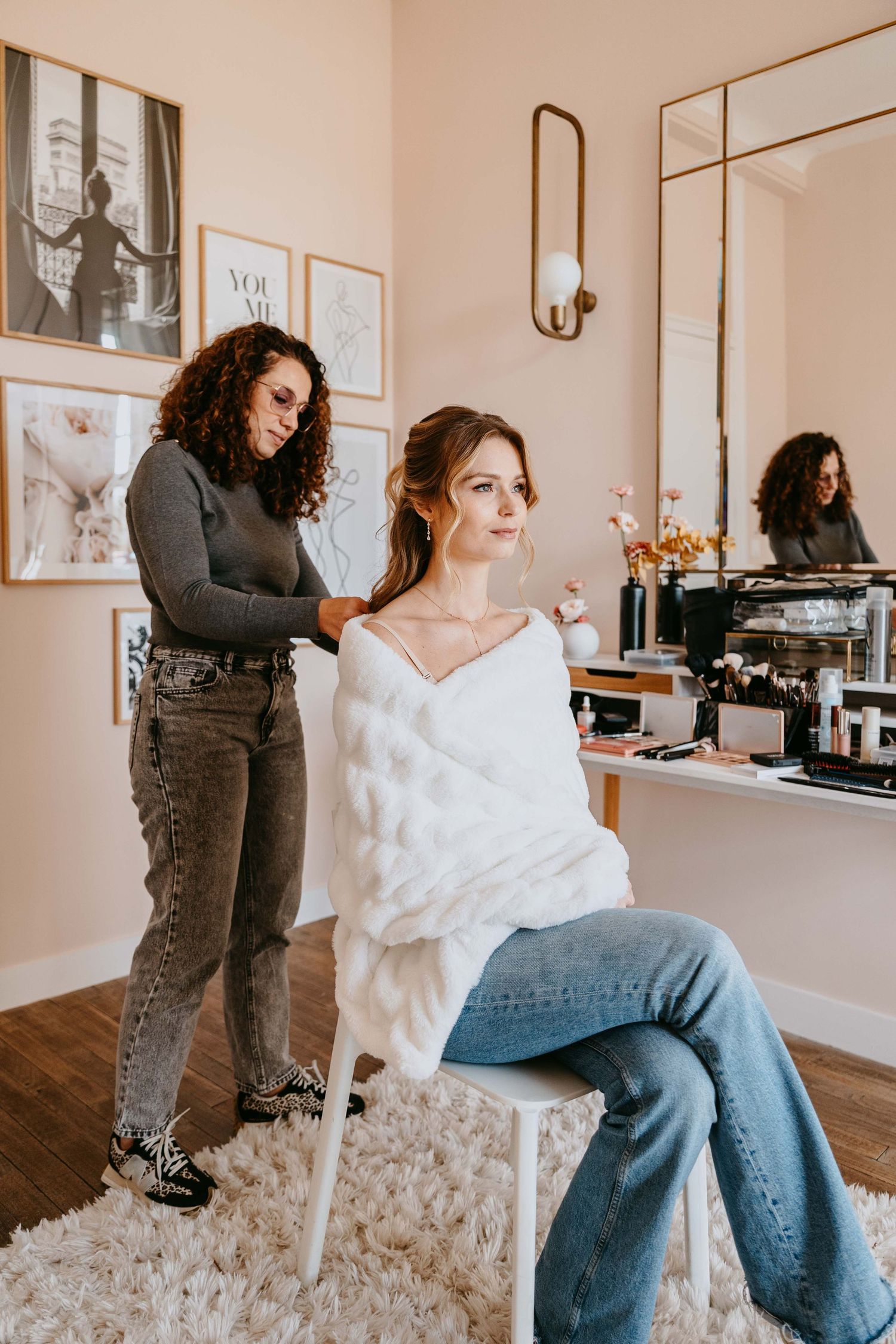 Curly-haired stylist arranging hair of smiling woman in white fur wrap seated in chic salon with gallery wall.