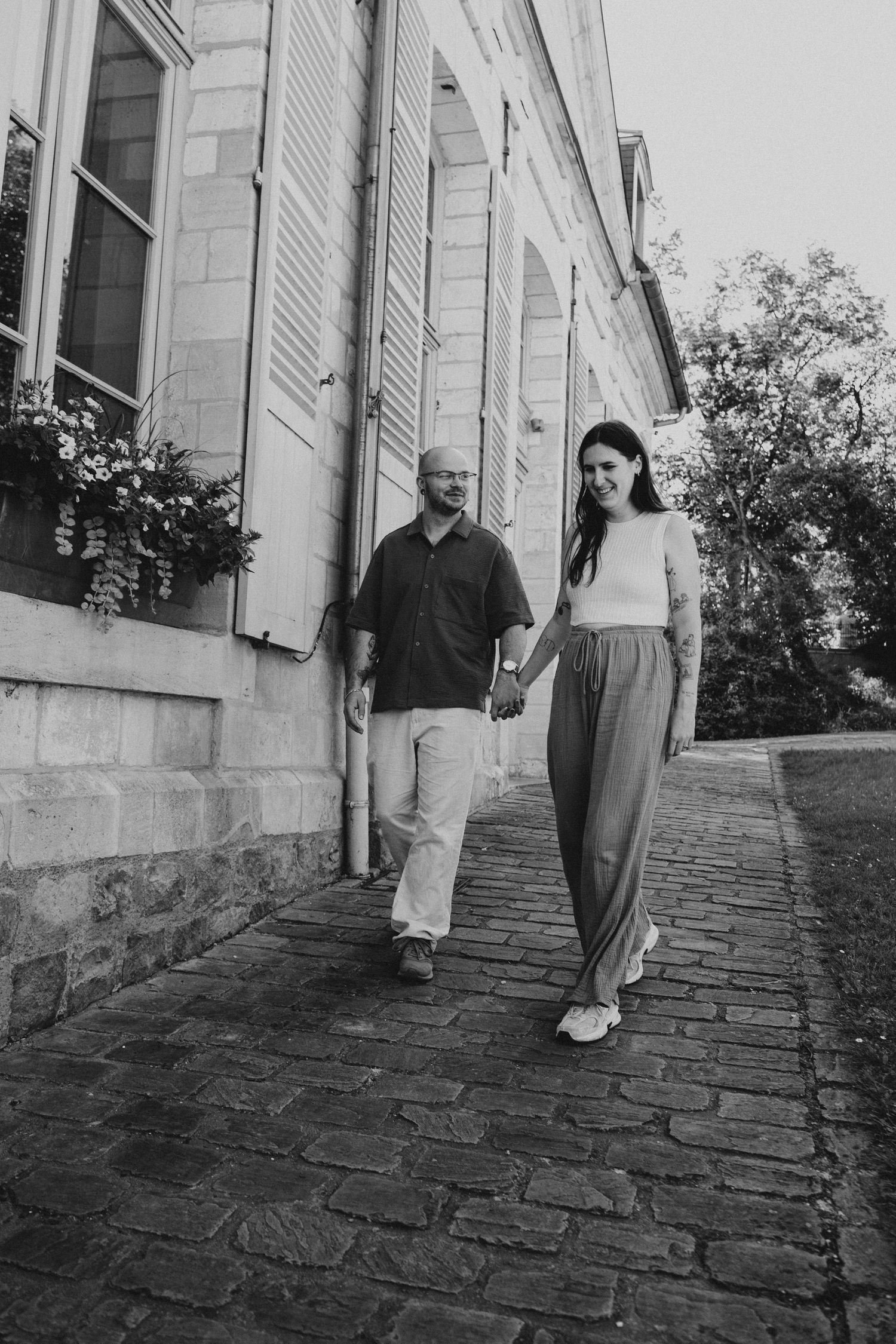 Black and white photo of a couple holding hands while walking on a cobblestone path beside a historic building.