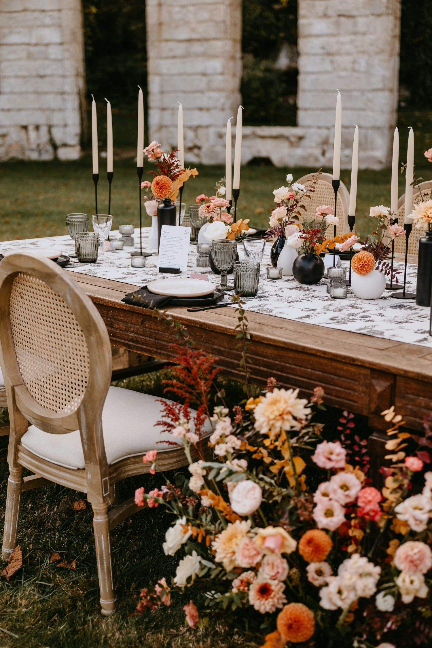 Elegant outdoor wedding tablescape with tall white candles, orange and pink florals, wooden farm table, and rattan chairs.